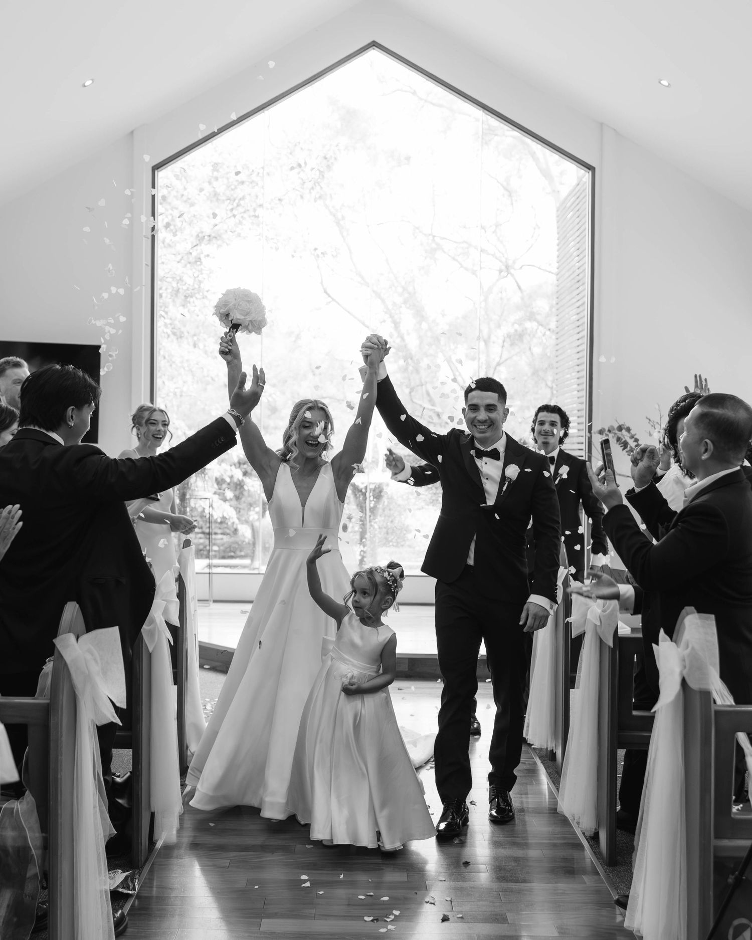 Newlyweds celebrate joyfully walking down aisle with flower girl and guests cheering in stunning chapel.