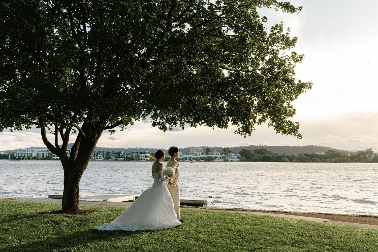Bride and groom stand beneath a large tree by a serene lakeside at sunset in their elegant wedding attire.