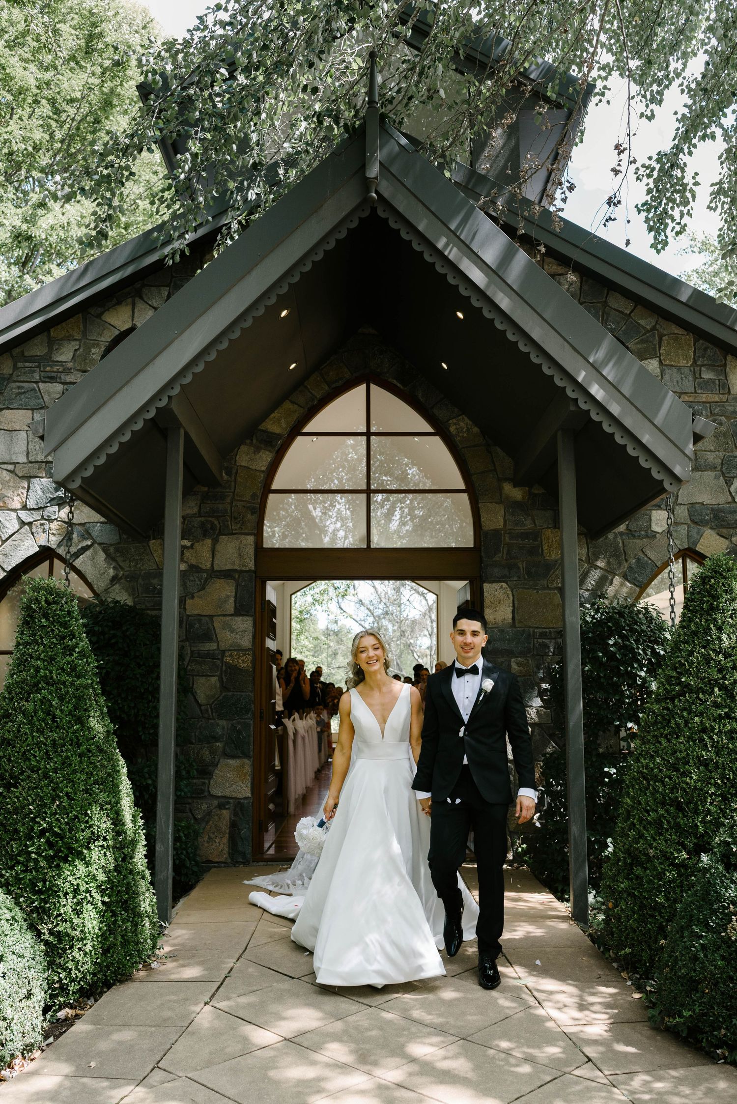 Bride and groom exit a charming stone chapel with arched windows, surrounded by lush greenery on their wedding day.