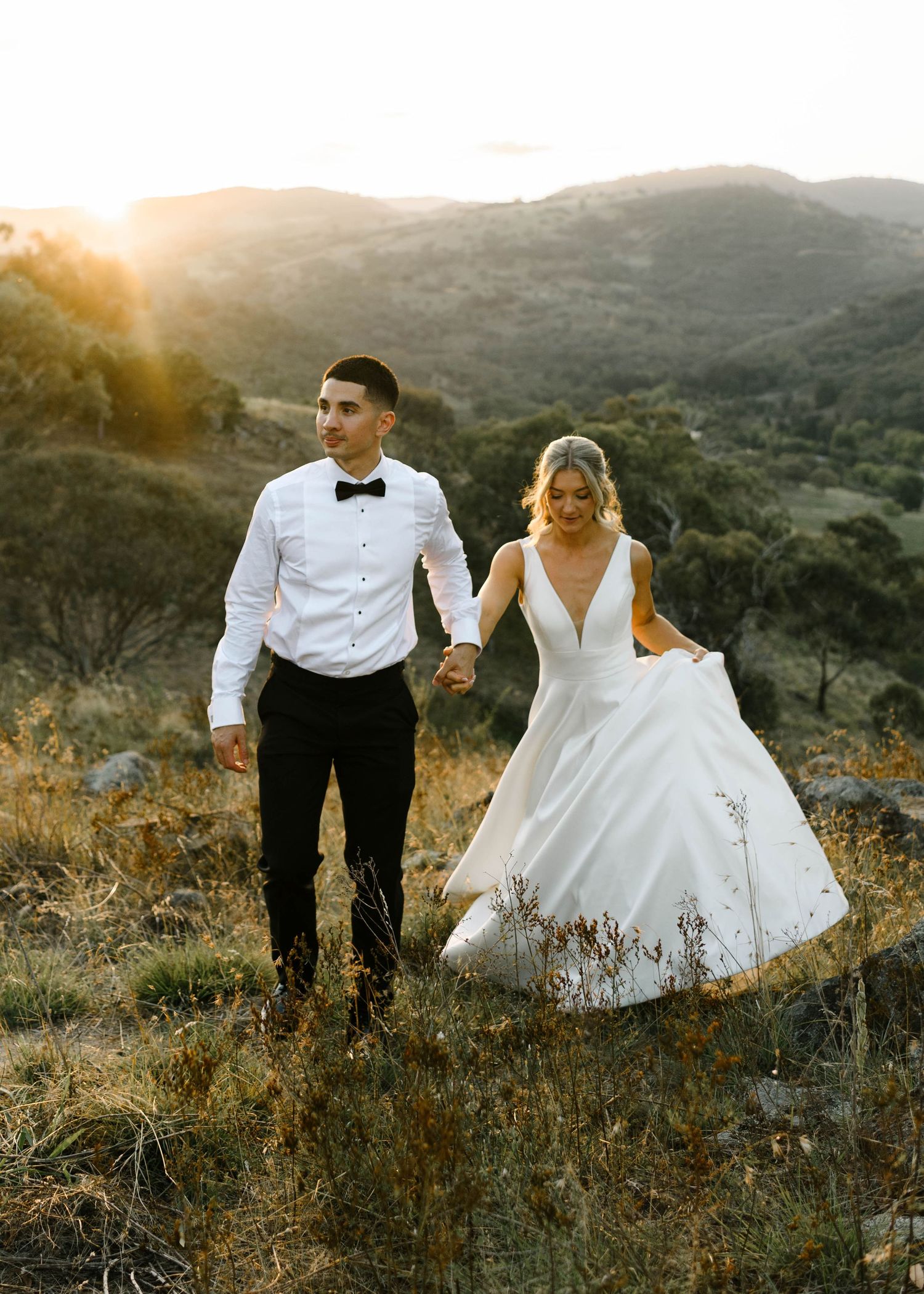 Bride and groom holding hands at golden sunset in rolling hills, bride in elegant white gown, groom in white shirt and bow tie.