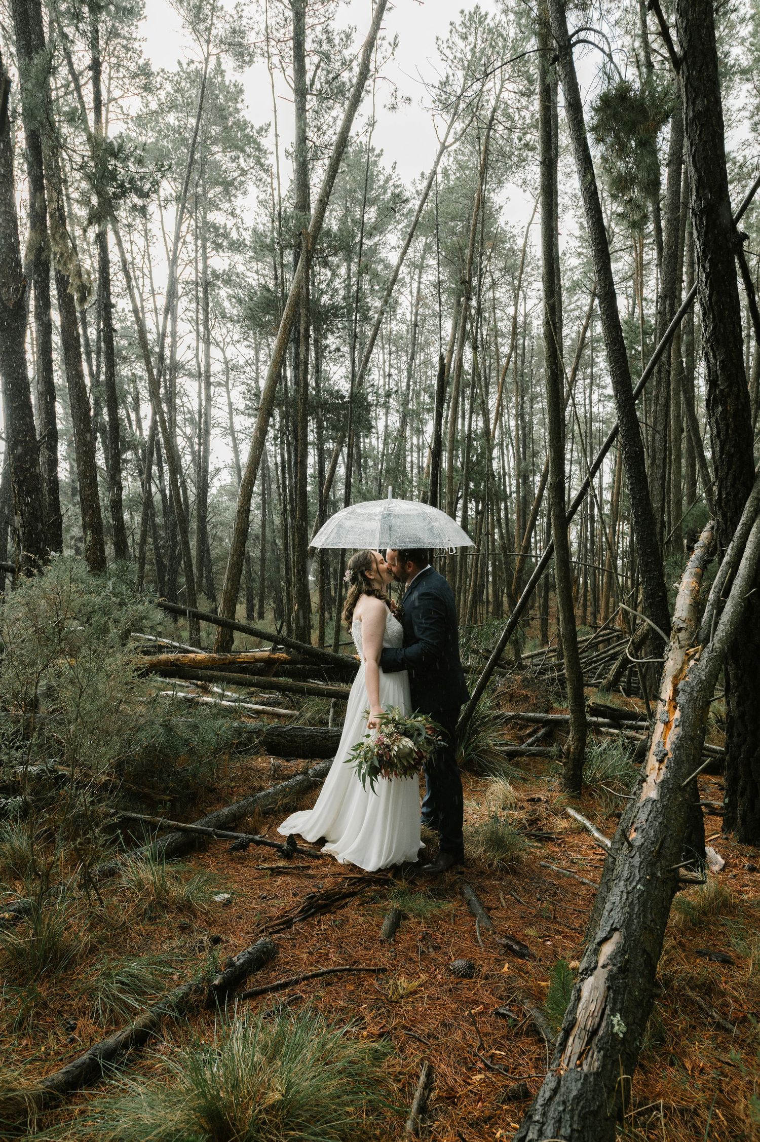 A bride and groom kiss under a clear umbrella in a misty forest, bride holding a bouquet in a white gown.