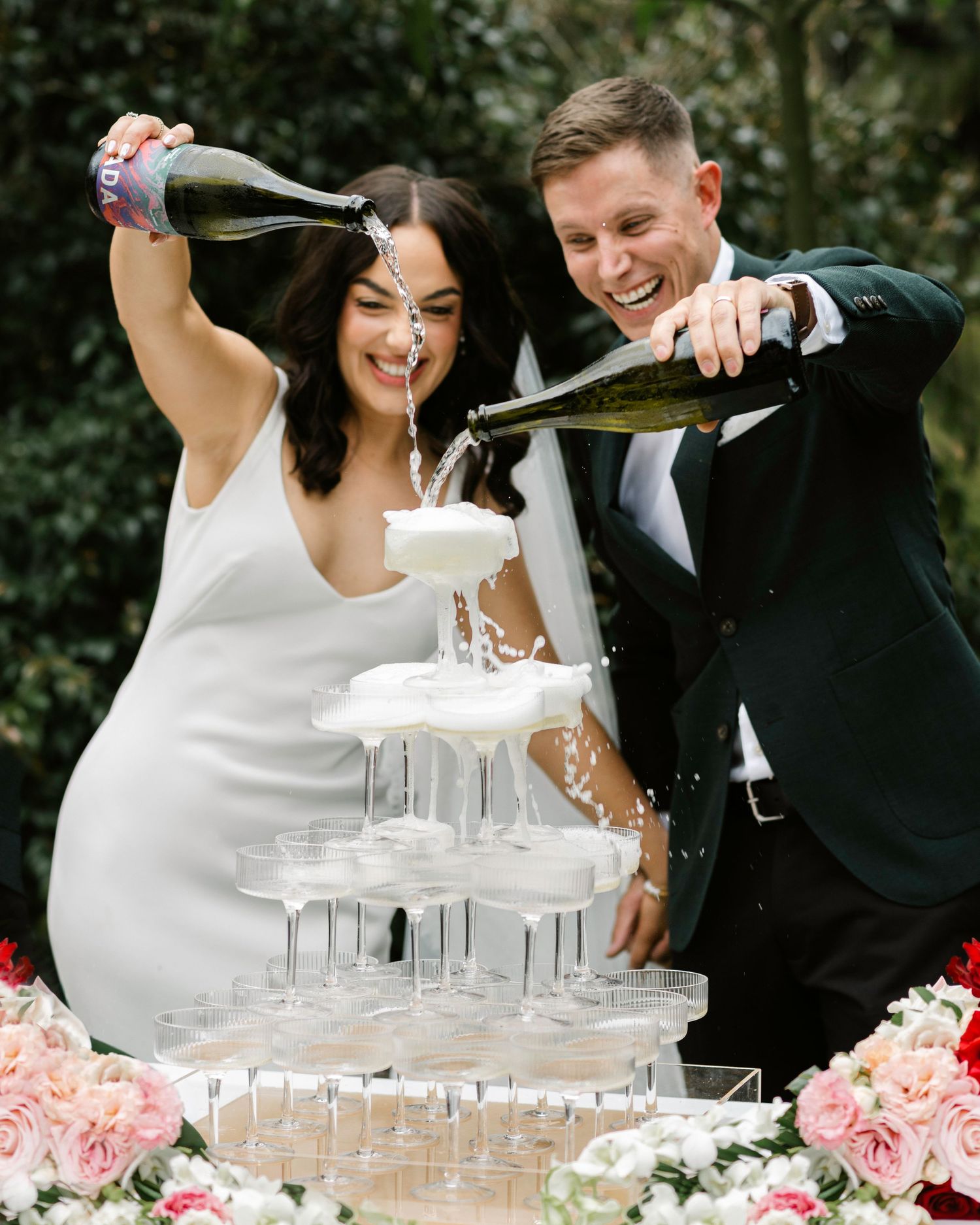 Bride and groom joyfully pouring champagne into a tiered glass tower surrounded by pink floral arrangements at their wedding.