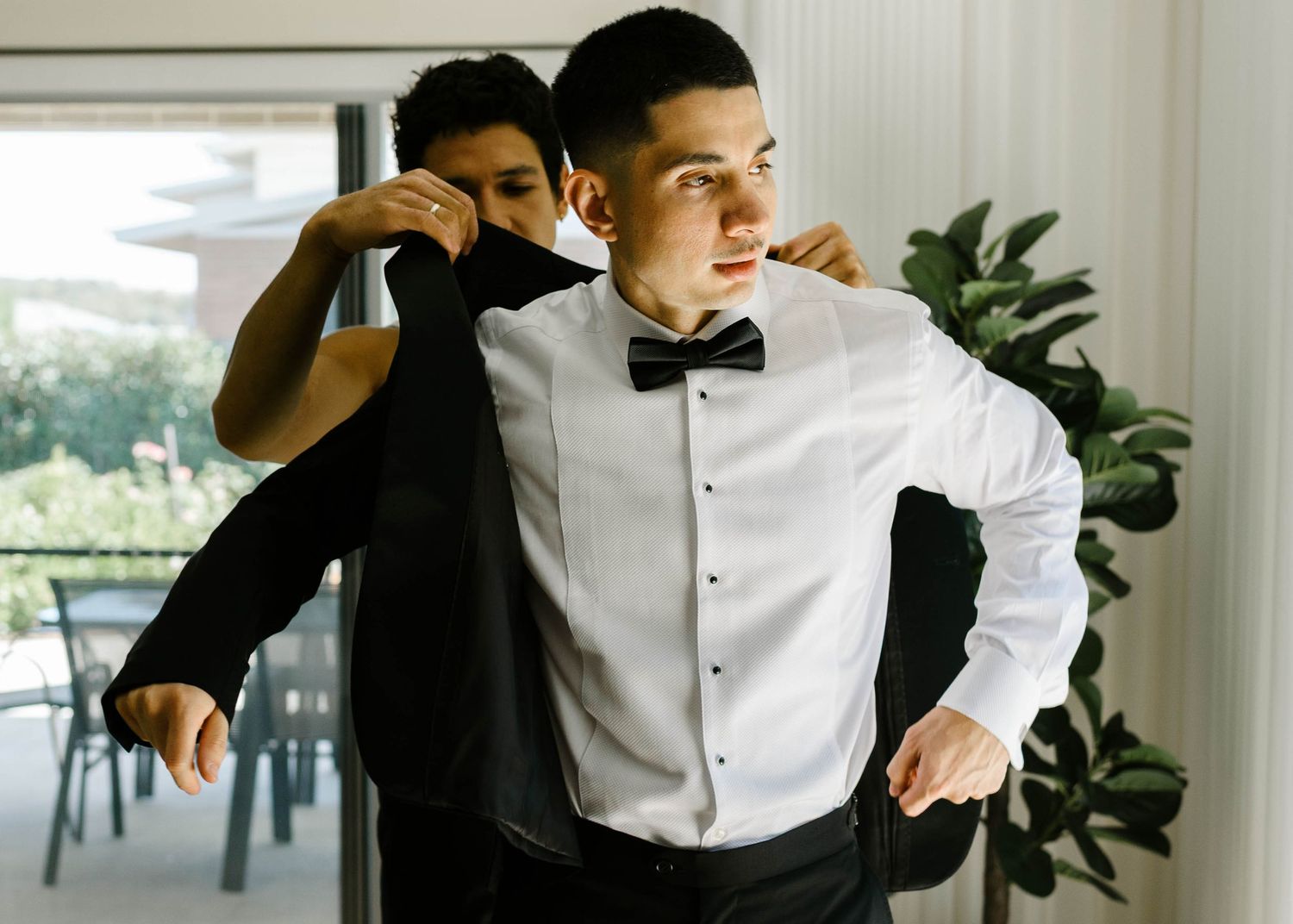 A young man in a tuxedo gets his bow tie adjusted by another person in a bright modern room.