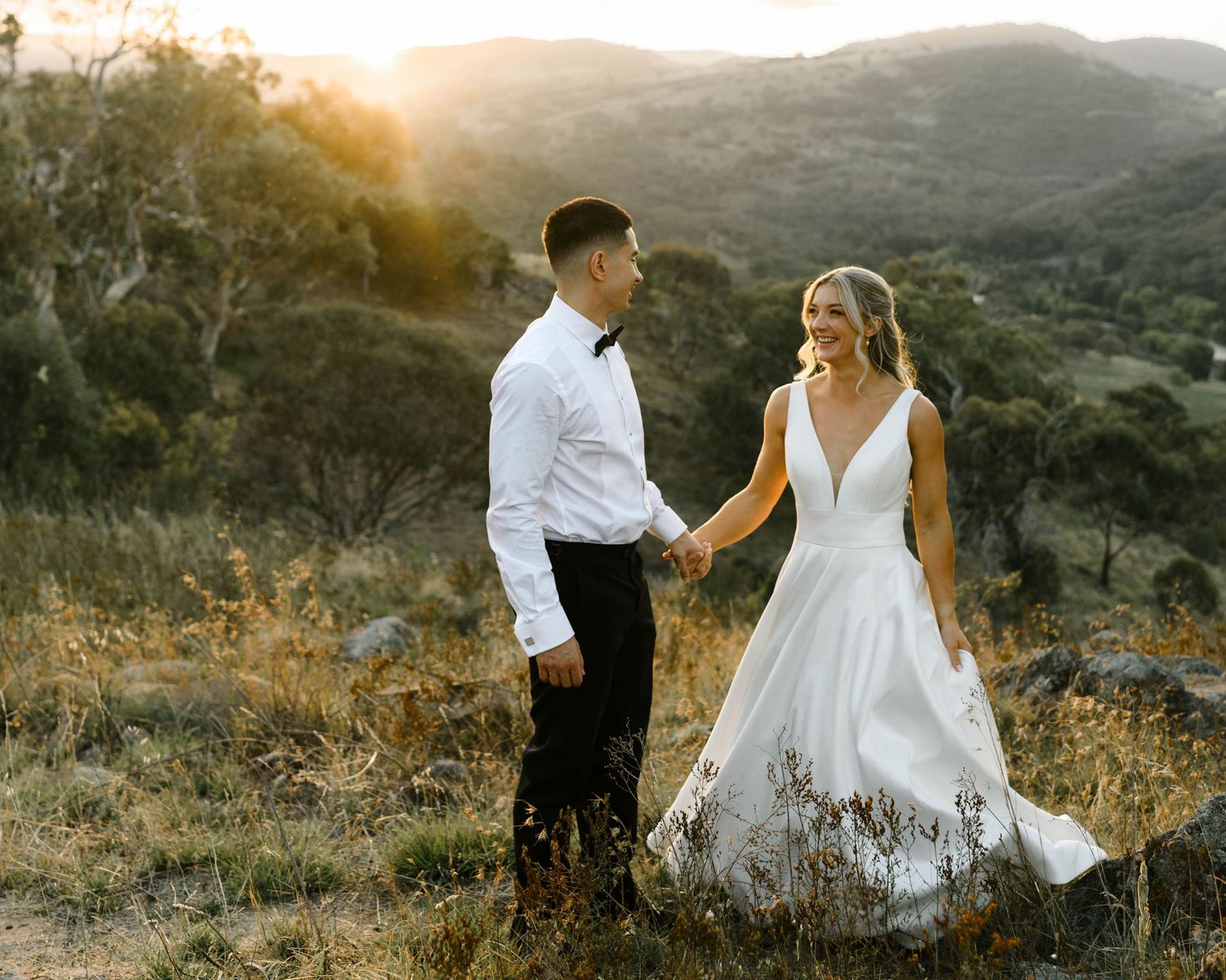 Couple in wedding attire holding hands at golden hour with scenic mountain landscape backdrop.