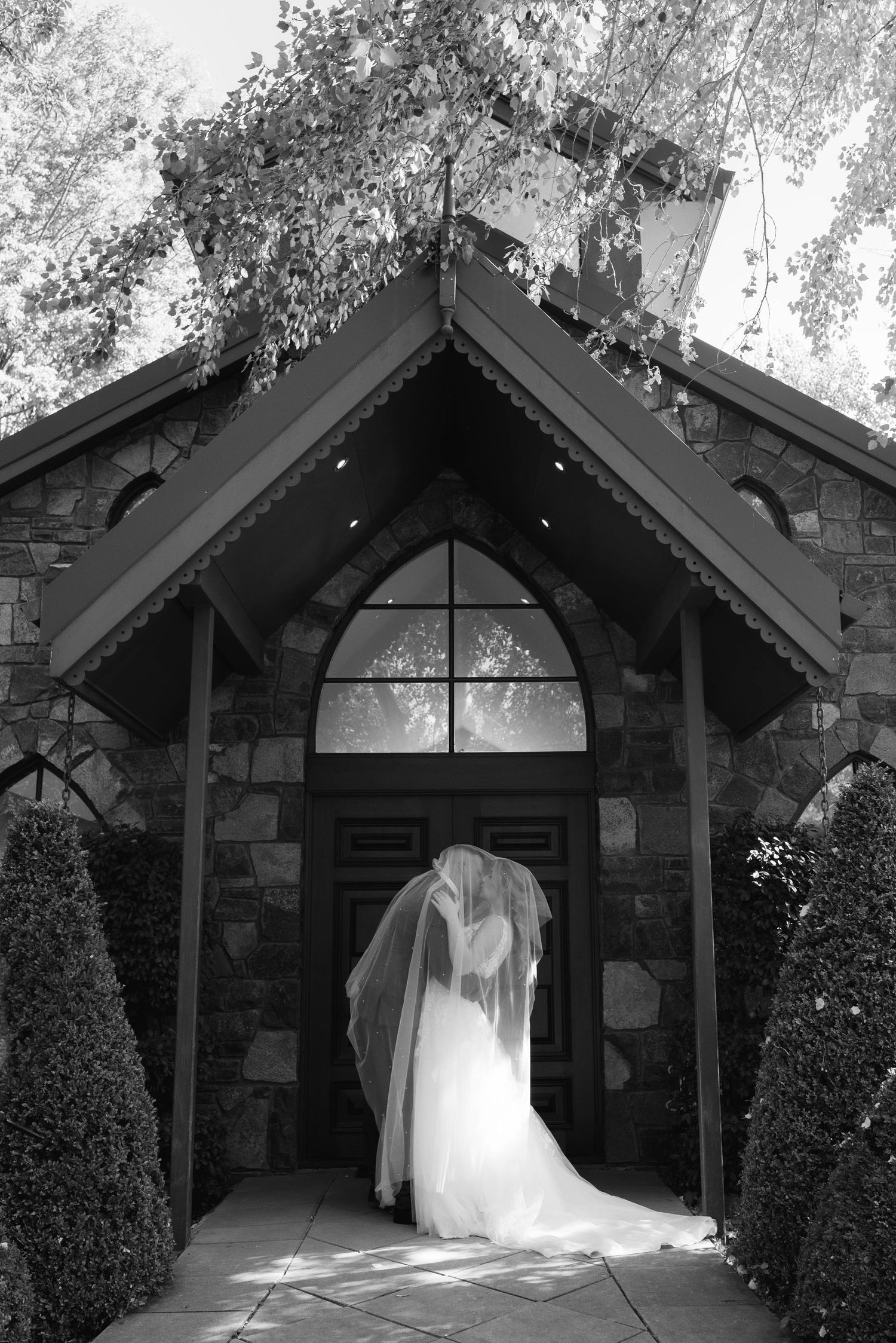A white wedding dress hangs elegantly in the doorway of a stone chapel with arched entrance and peaked roof.