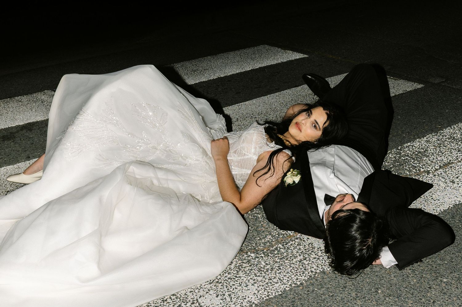 Bride in white gown and groom in tuxedo lying on a crosswalk at night in a romantic wedding photo.