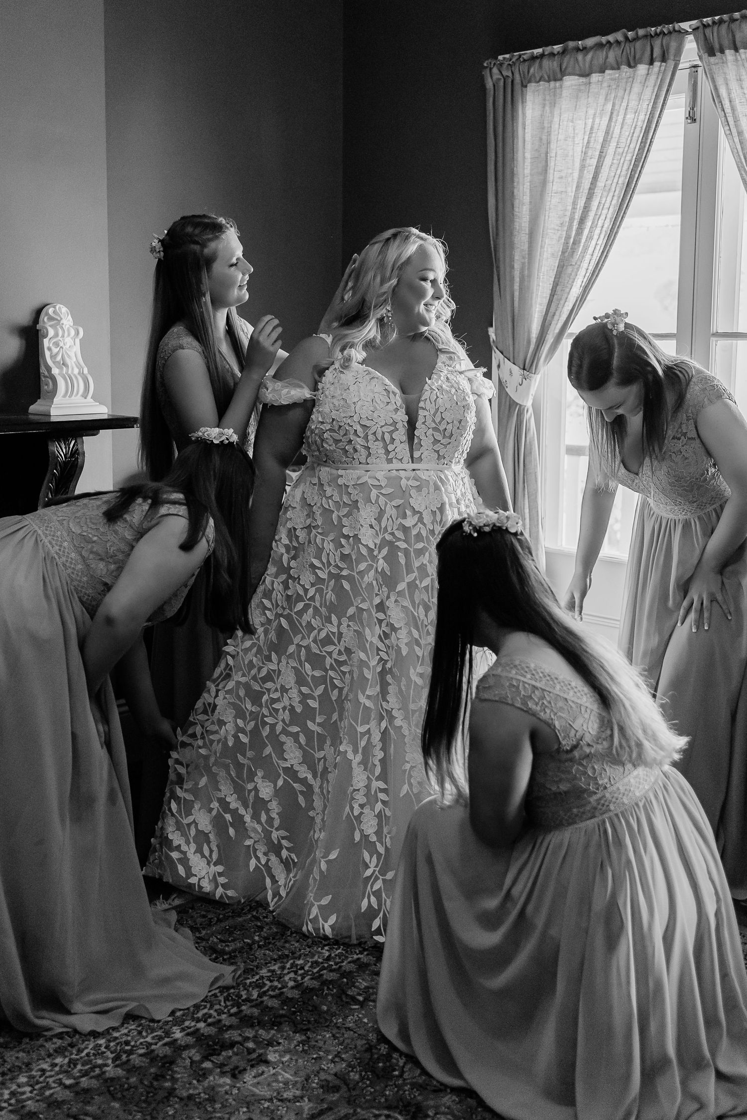 Black and white photo of a bride in a lace gown being helped by bridesmaids near a window before her wedding.