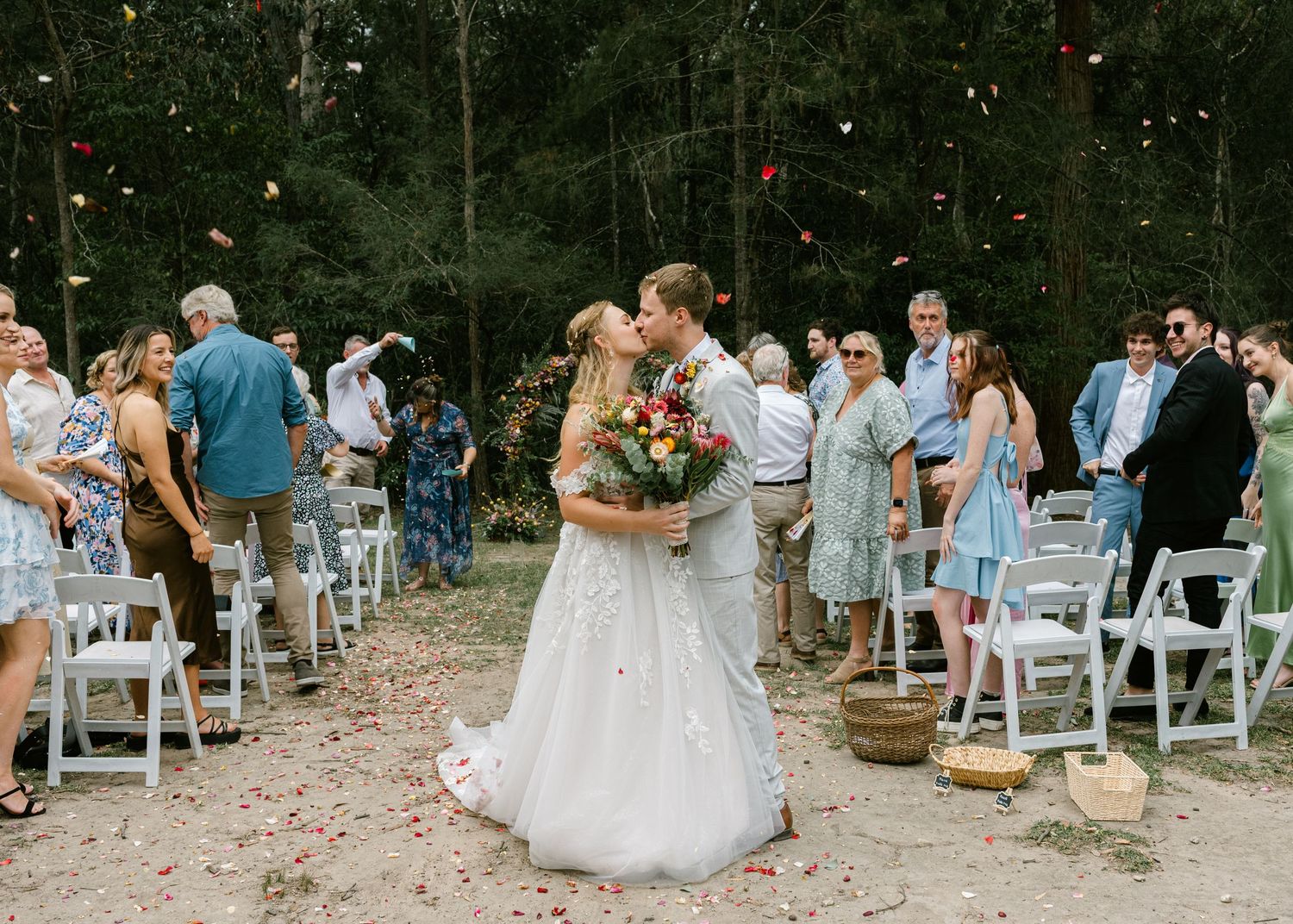 Bride and groom share first kiss at outdoor wedding ceremony while guests toss flower petals in forest setting.