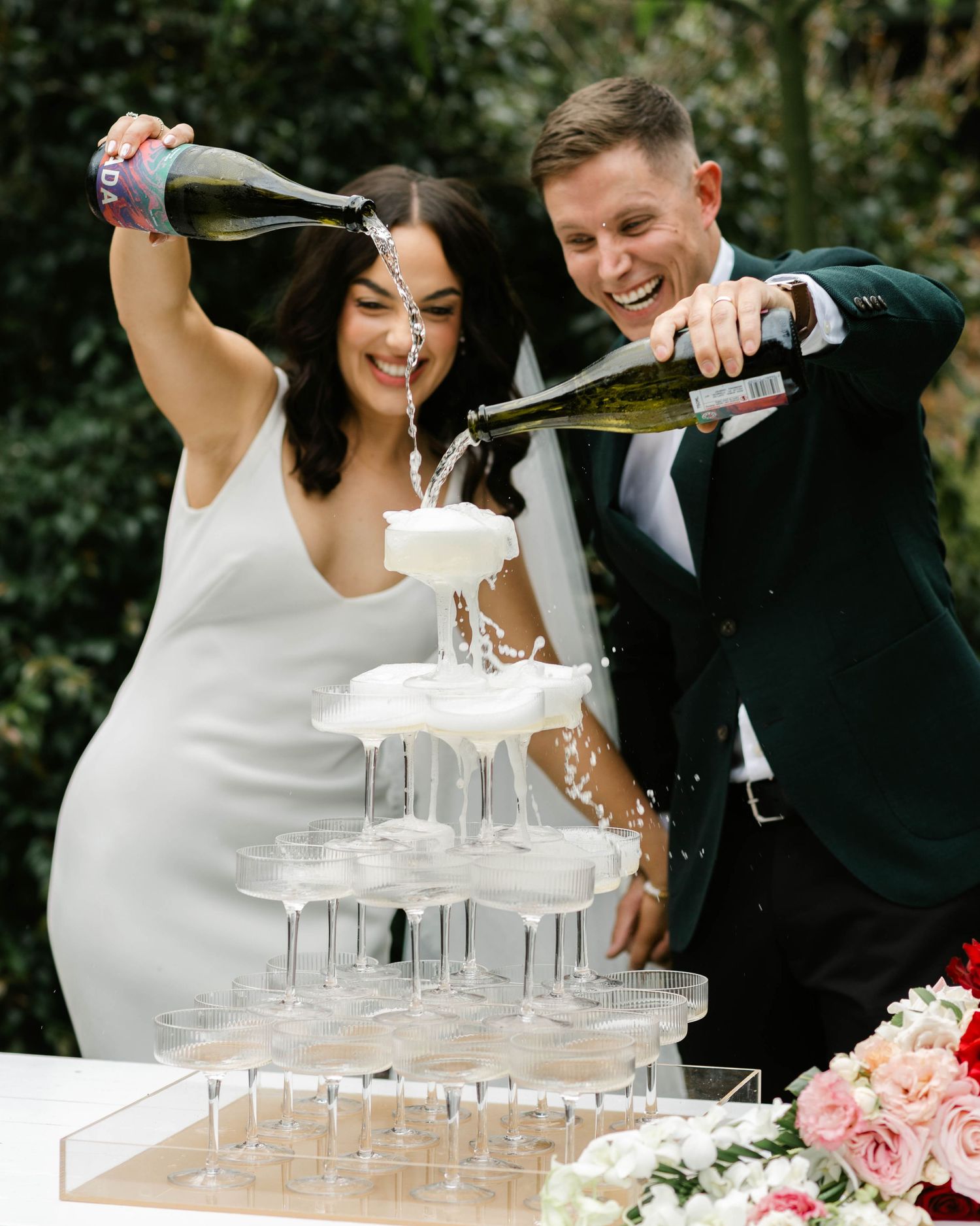 A couple pours champagne into a tower of glasses at their wedding celebration outdoors.