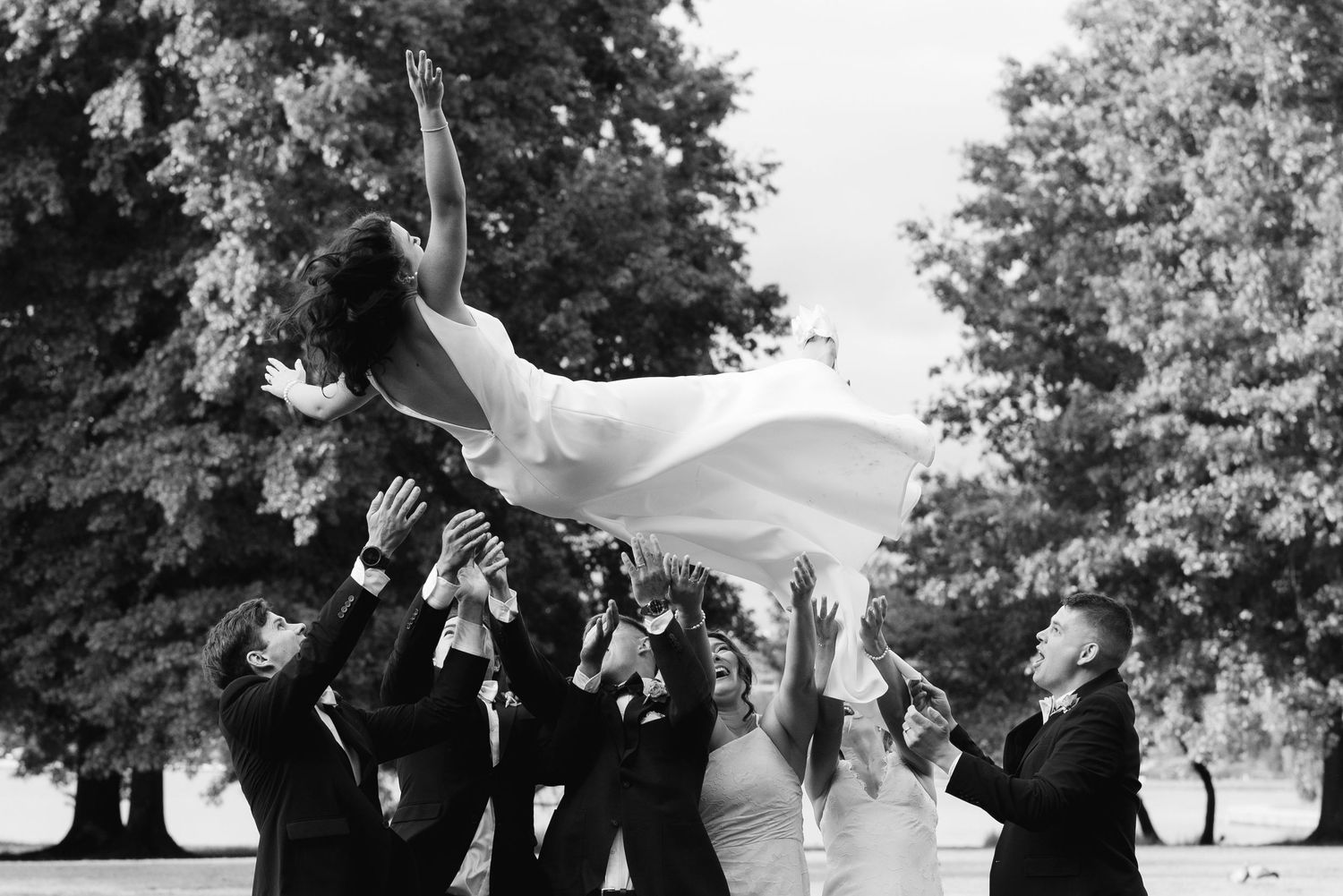 Bridal party joyfully tossing bride into the air outdoors in a black and white wedding photo.