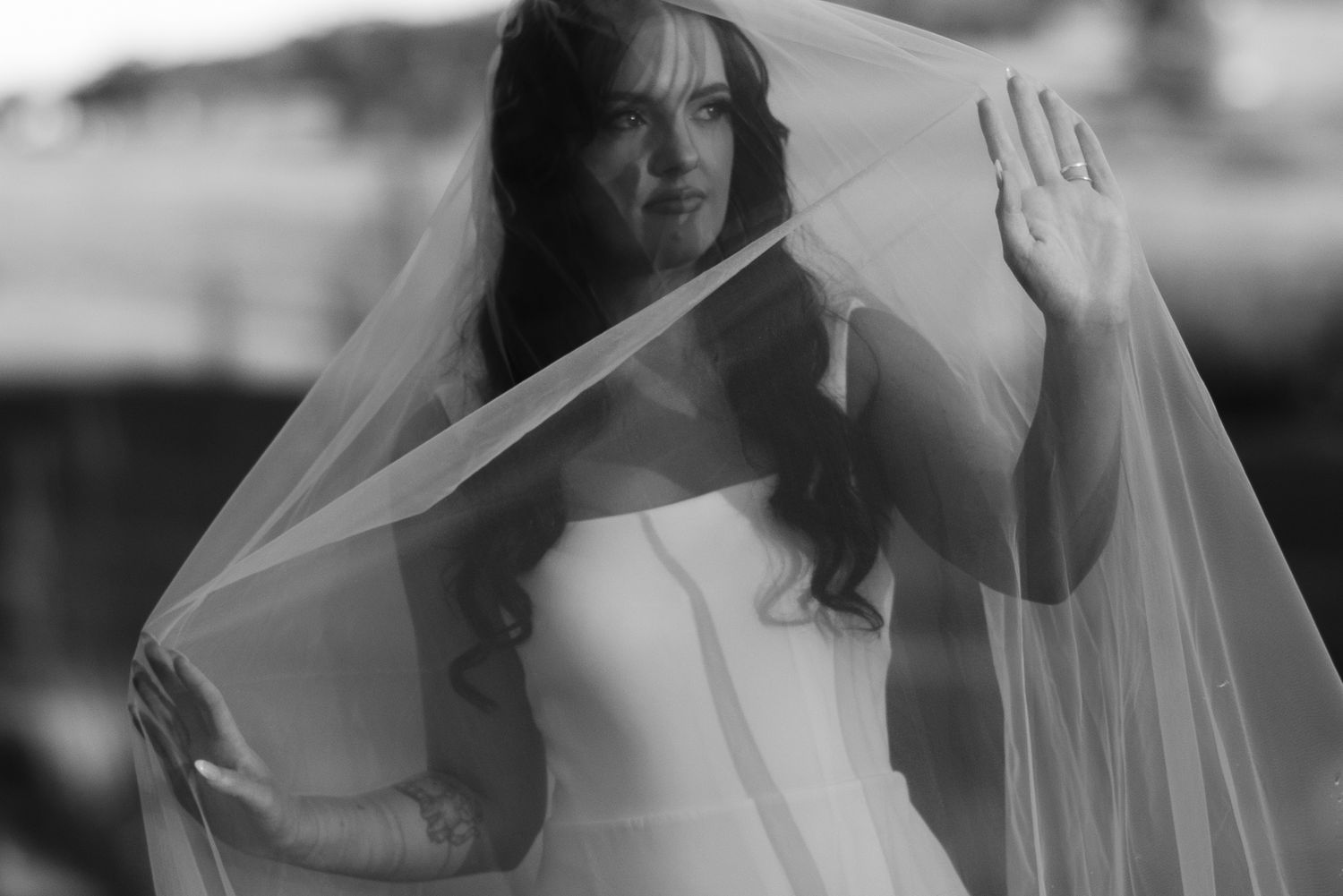 Black and white photo of a bride in a white gown lifting her veil, looking up gracefully at an outdoor wedding.