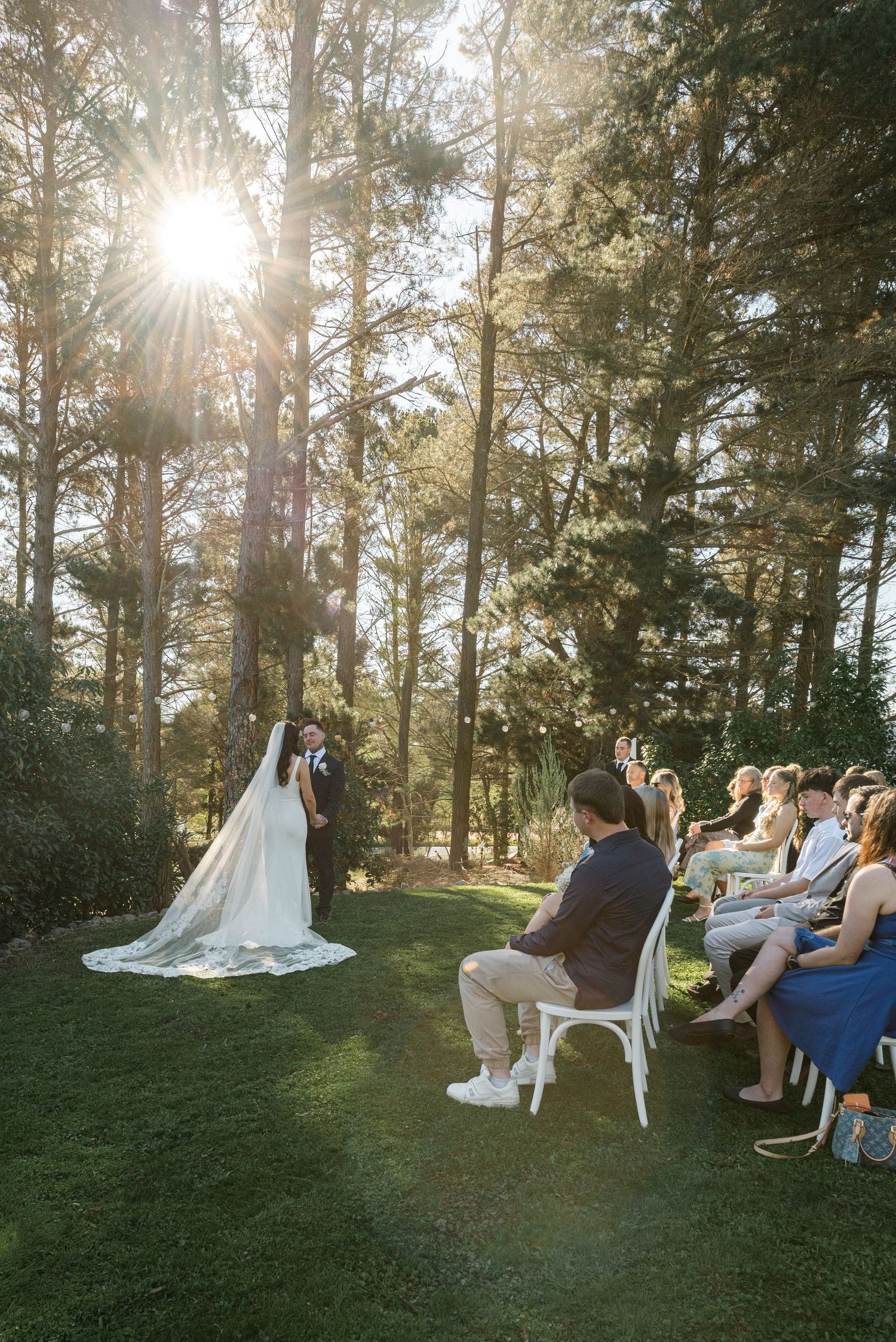 Outdoor wedding ceremony in a sunlit forest, bride and groom kissing at the altar as guests watch from white chairs.