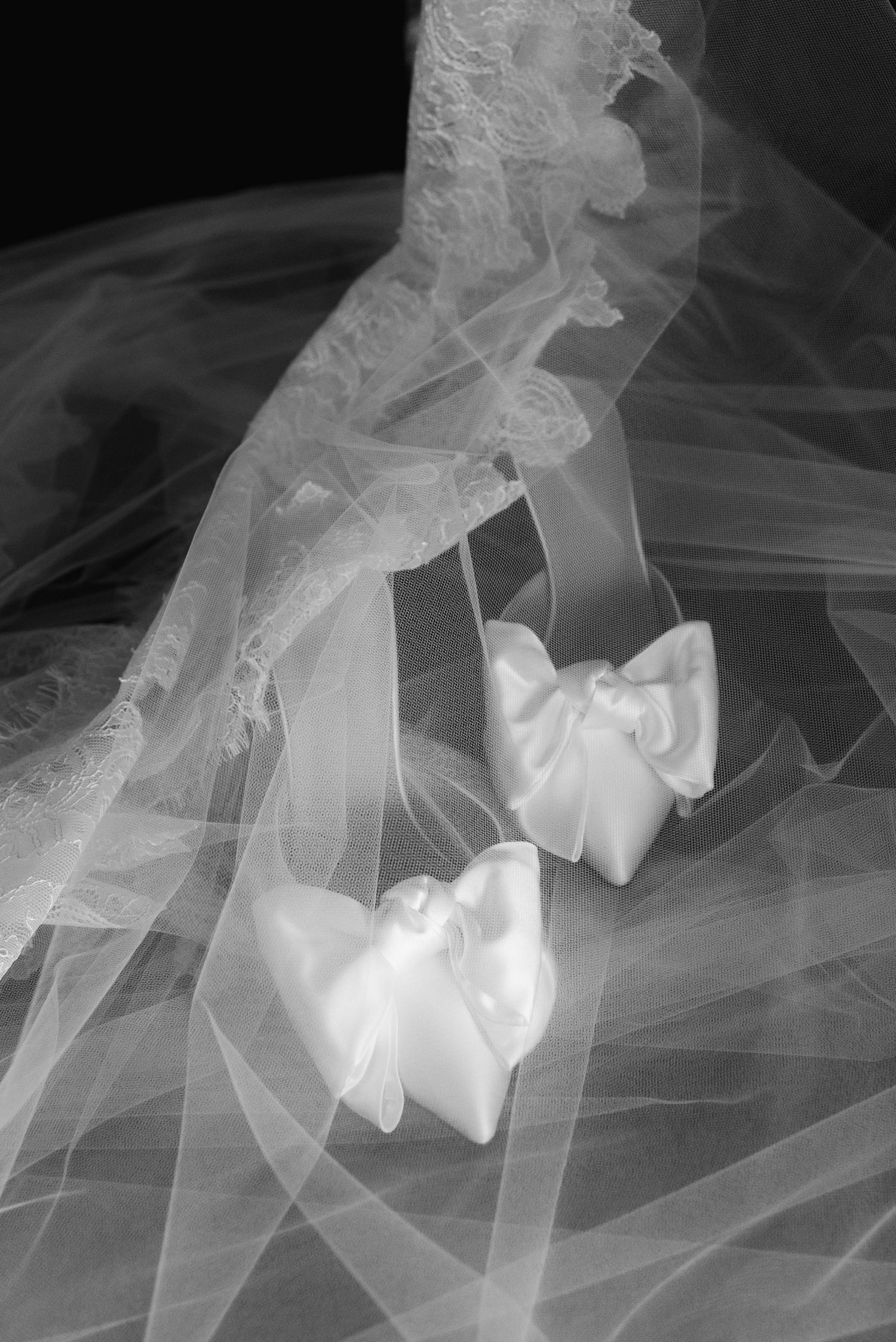 Black and white photo of elegant satin bridal shoes with bows resting on a flowing wedding veil and lace.