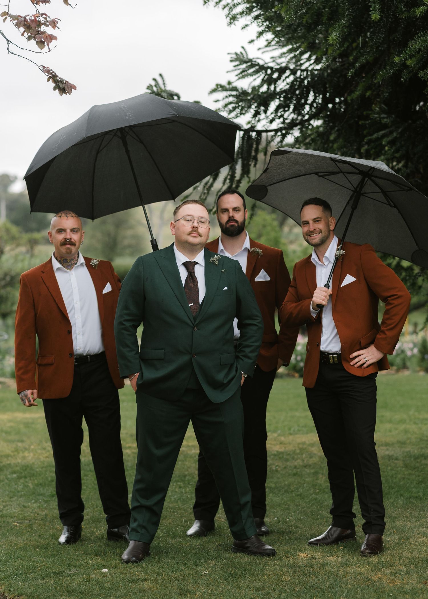 Groom in green suit stands with three groomsmen in rust blazers holding black umbrellas outdoors.