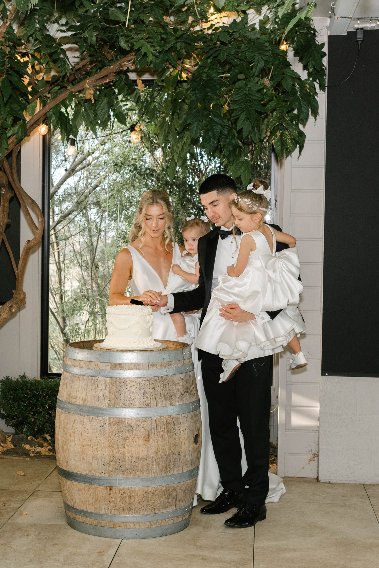 Bride and groom cutting wedding cake on barrel with two young children, surrounded by lush greenery and string lights.