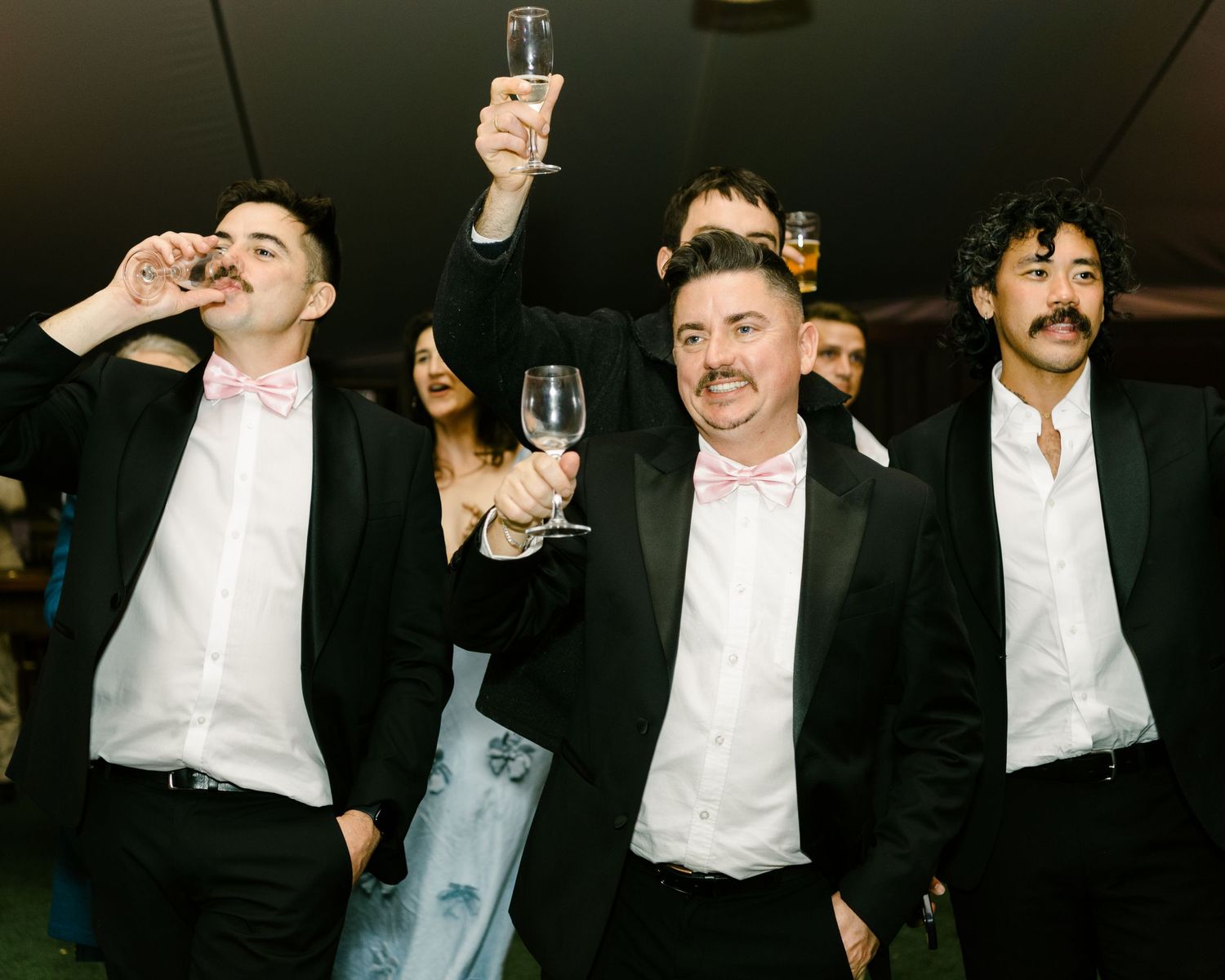 Three men in black tuxedos with pink bow ties toasting with champagne glasses at a formal event.