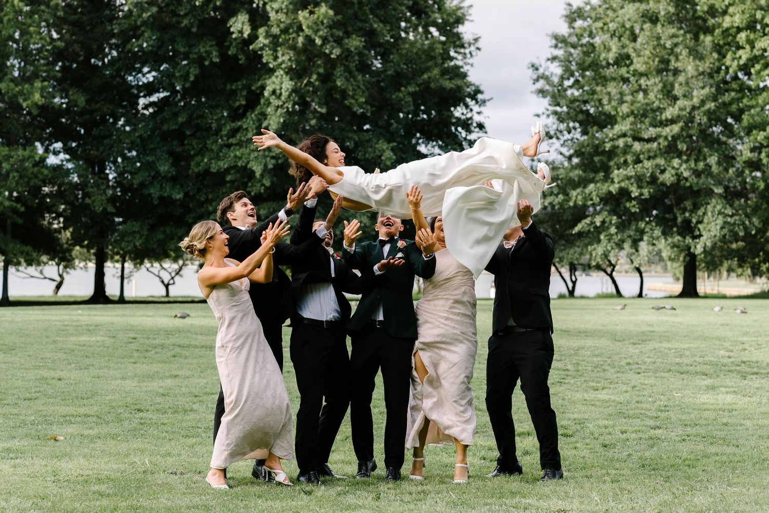 Wedding party members jump and pose playfully together on a grassy lawn during outdoor celebration photos.