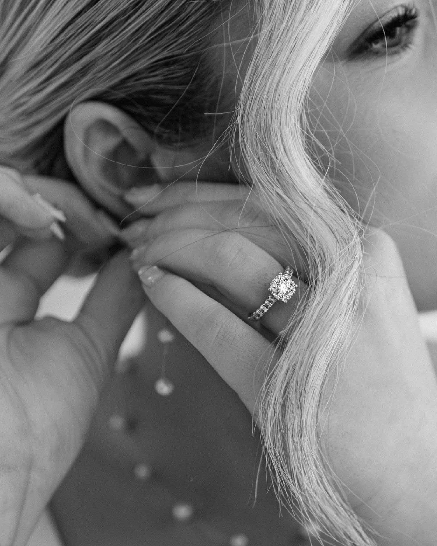 Black and white photo of a woman wearing a diamond engagement ring while tucking her blonde hair behind her ear.