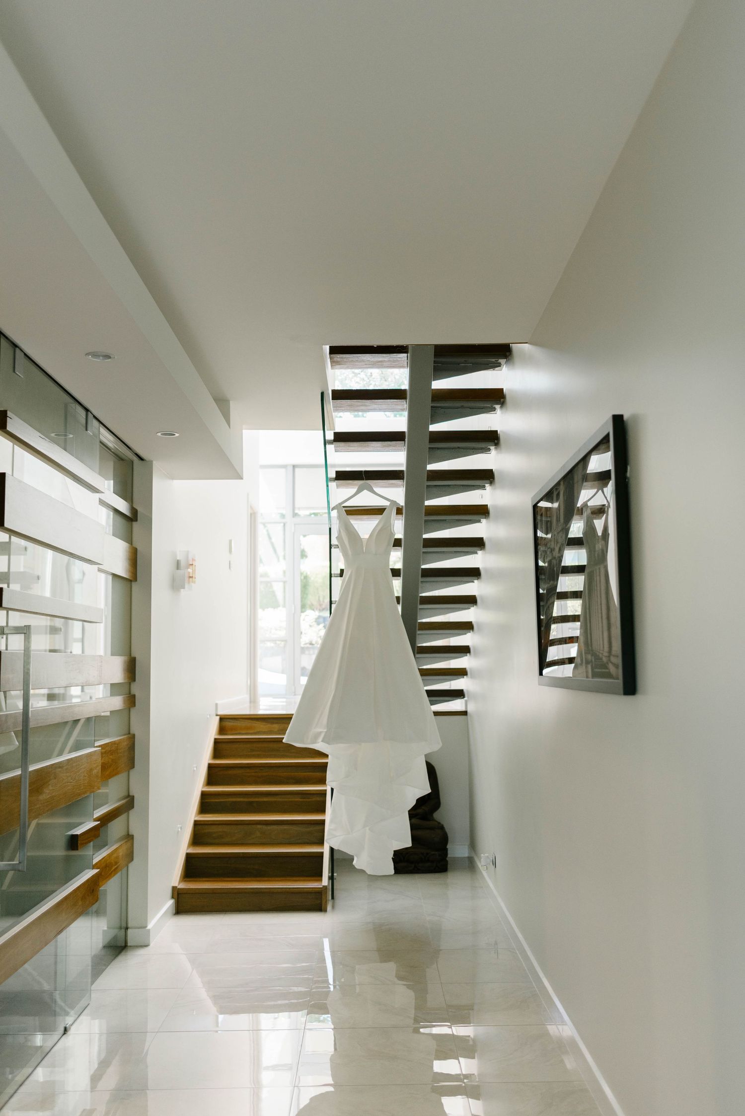 White wedding dress hanging on staircase in modern home with glass railings and wooden steps.