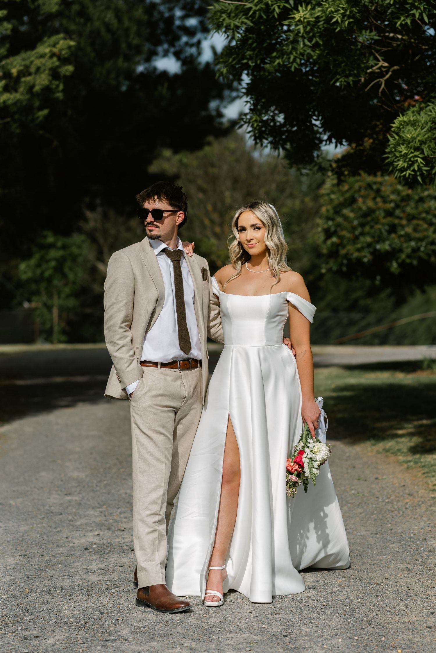Bride in white off-shoulder gown with slit and groom in tan suit walk together outdoors on a sunny wedding day.