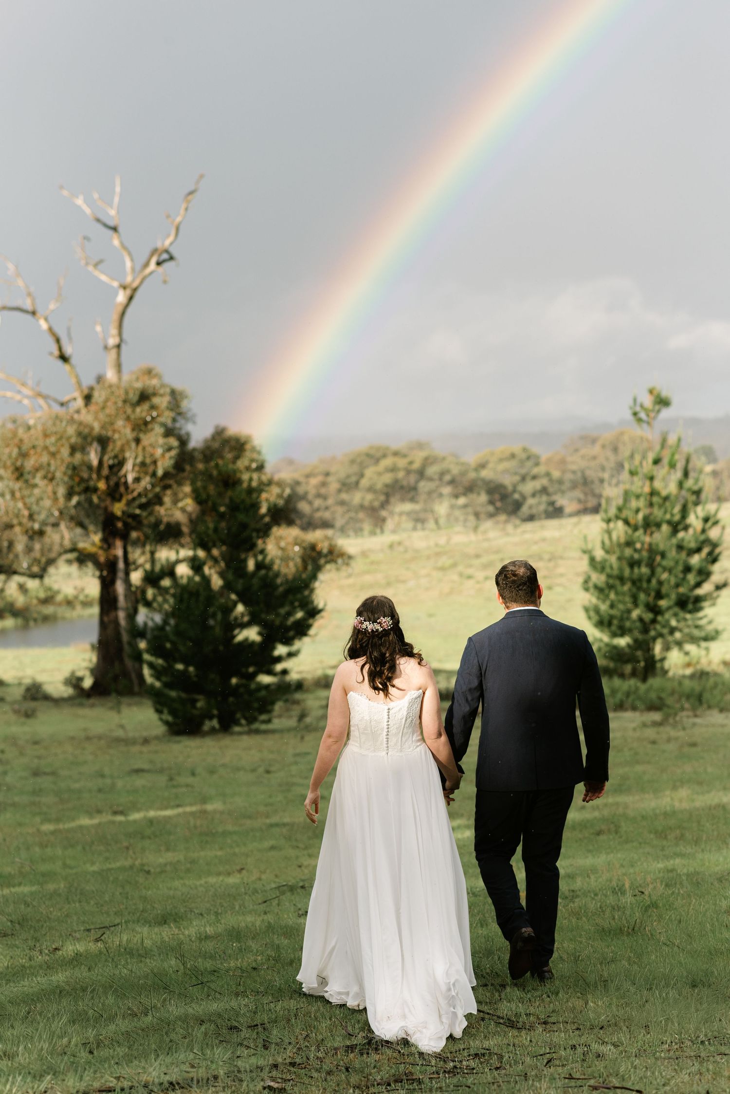 Bride and groom walk hand in hand across green field with stunning rainbow in background at outdoor wedding.