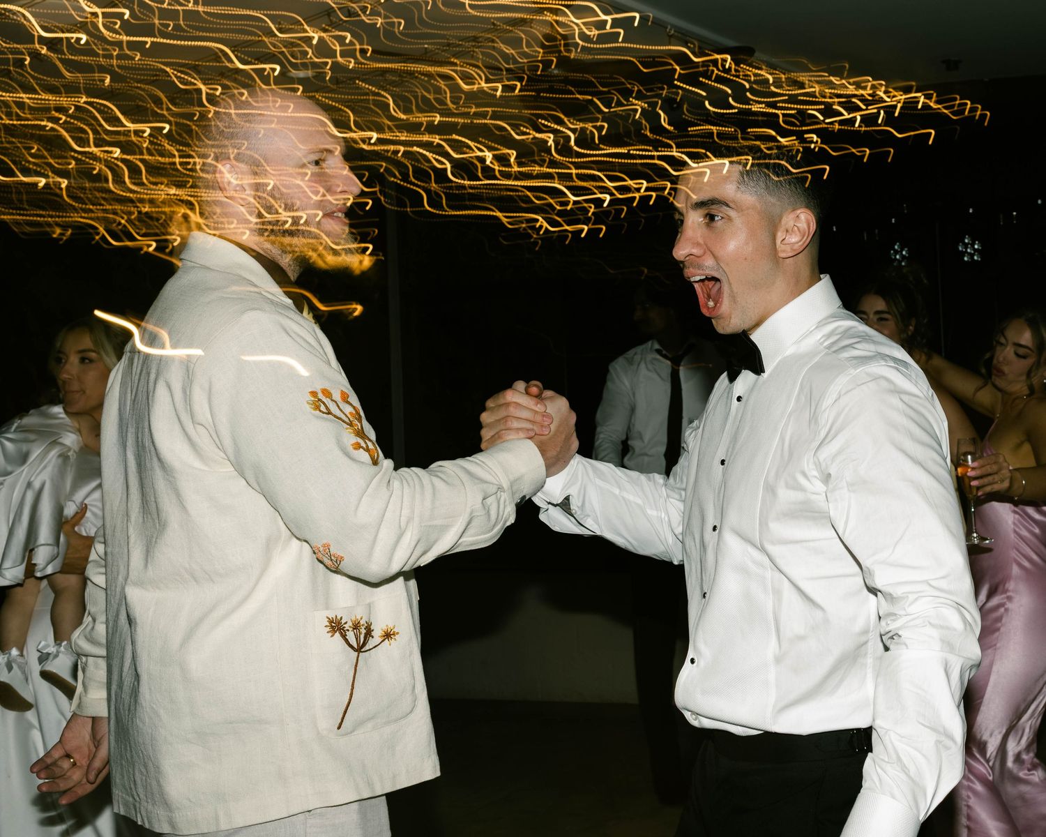 Two men in formal attire shaking hands and smiling at a dance event with bokeh light trails in background.