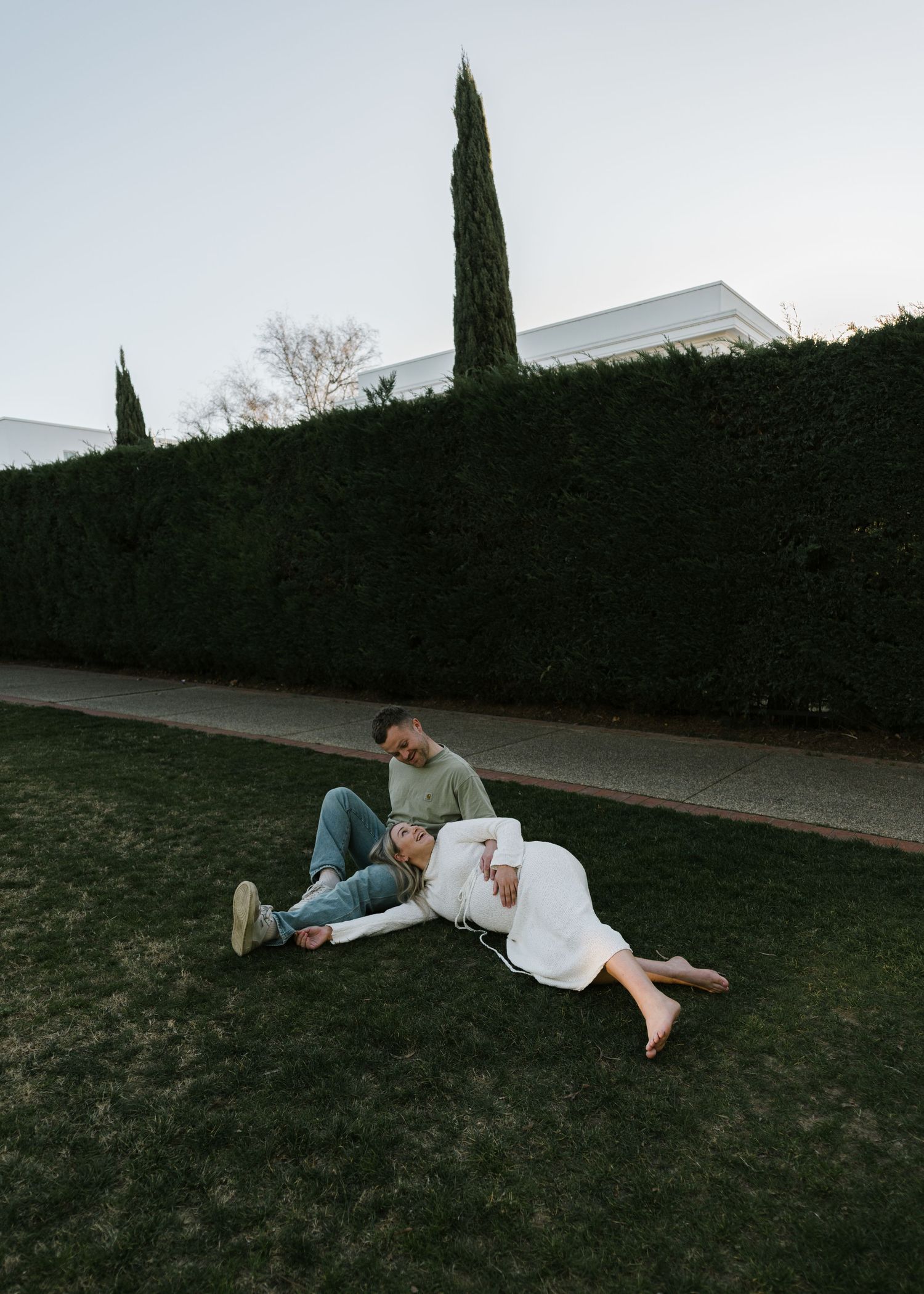 Couple relaxing on green lawn near tall hedges, woman in white dress leaning back on man in casual attire.