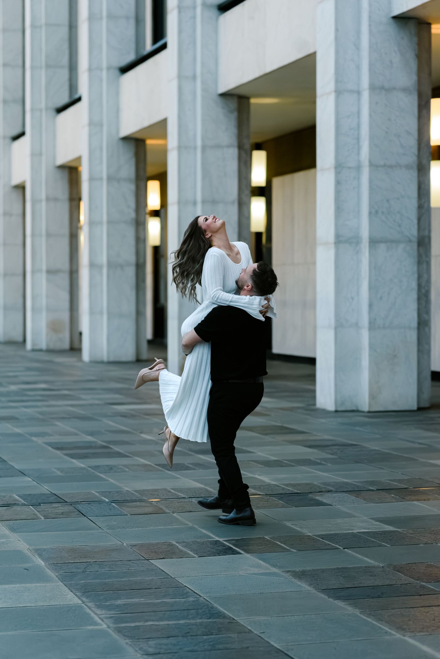 A man lifts a woman in a white dress as she laughs joyfully, posing between marble columns outdoors.