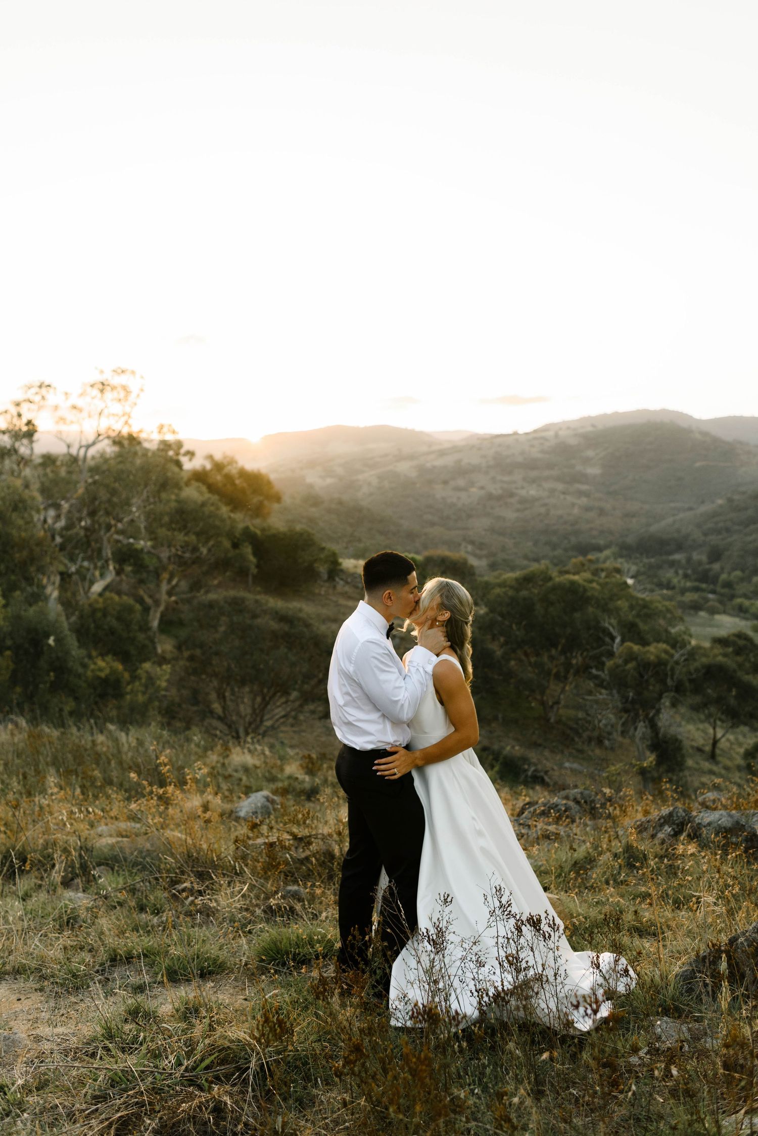 Bride and groom share romantic kiss at golden sunset in scenic Australian bush landscape.