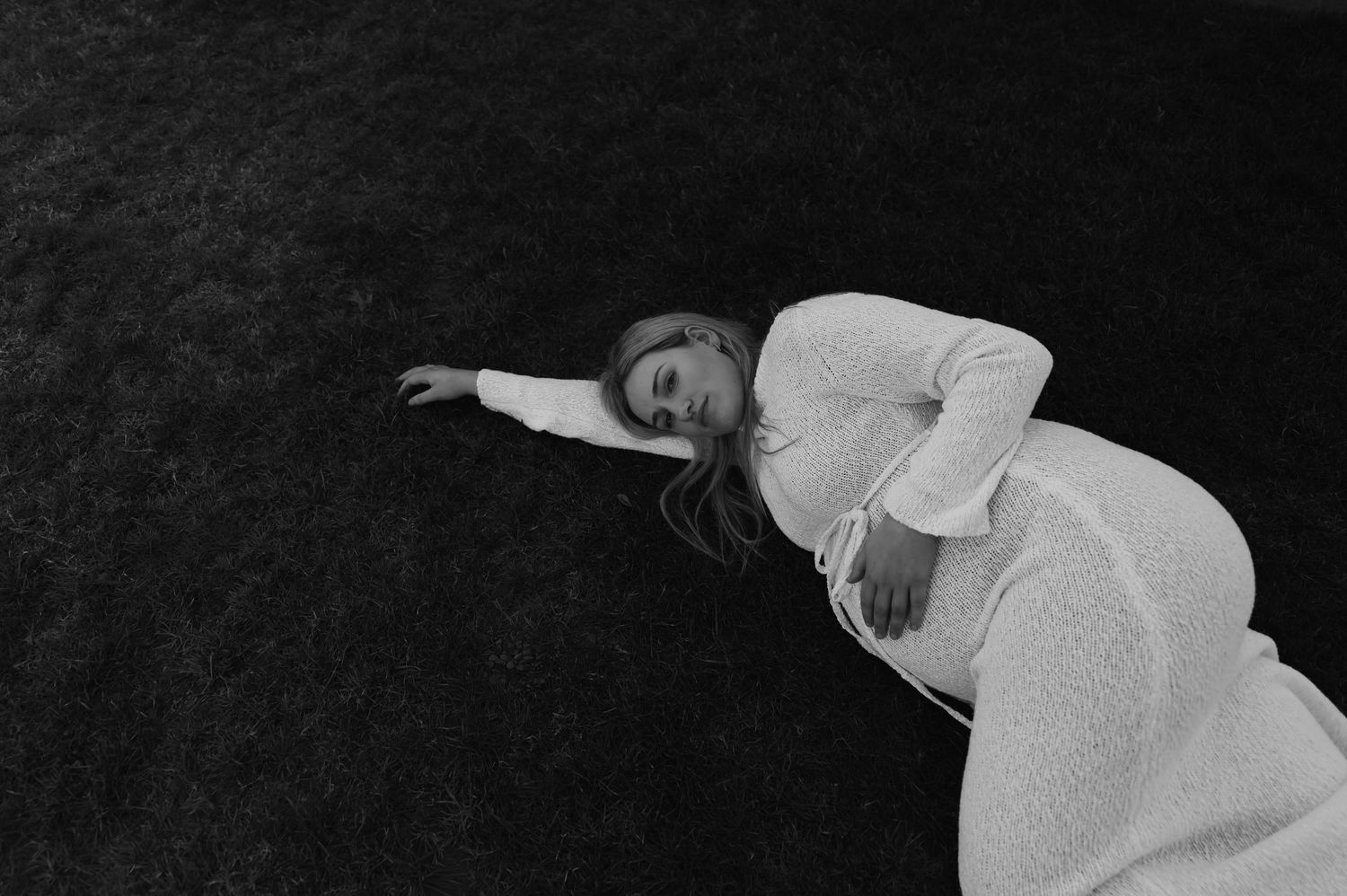 Black and white photo of a pregnant woman in white dress lying on grass, eyes closed, serene maternity portrait.