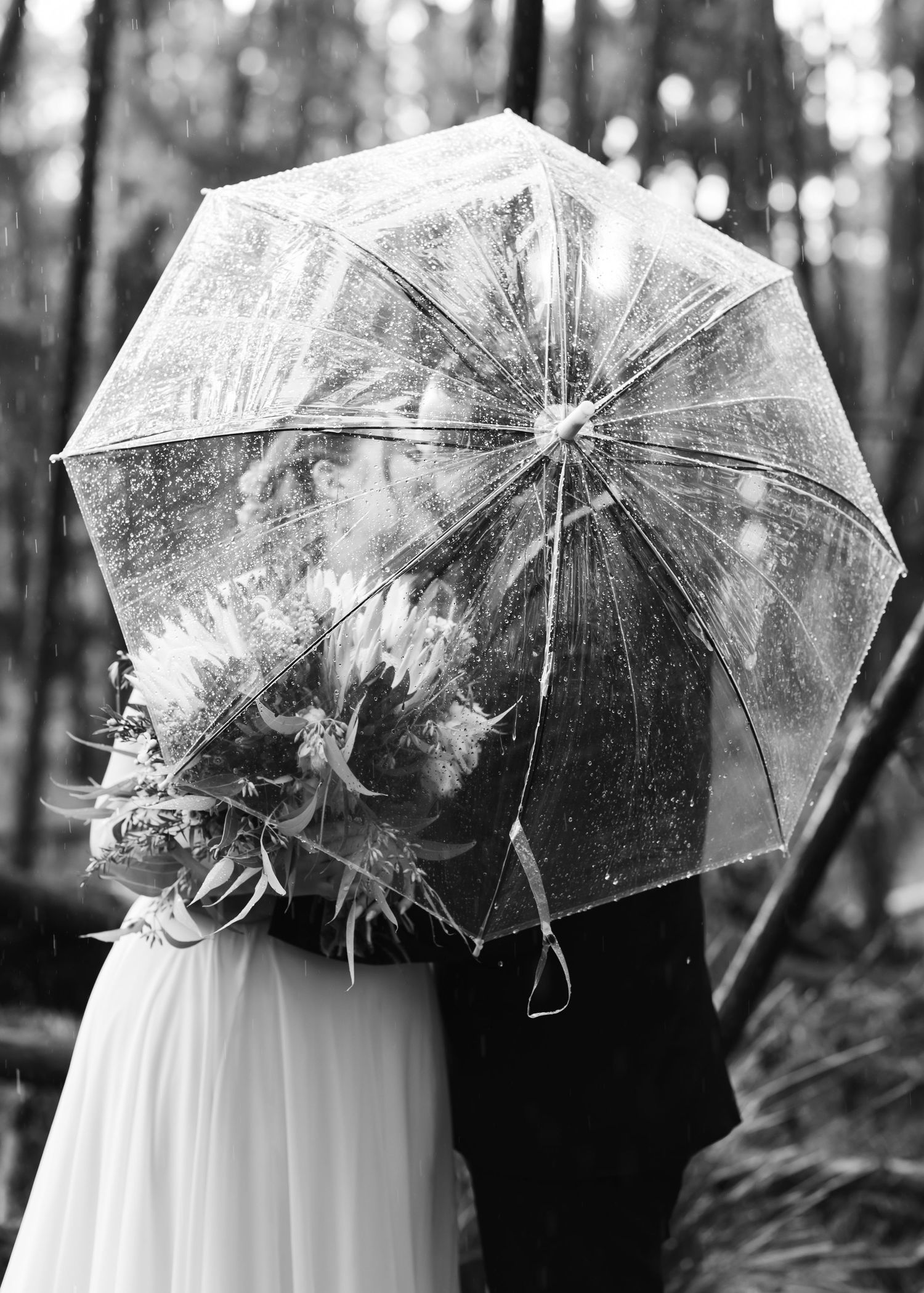 Black and white photo of a couple kissing under a clear umbrella in the rain on their wedding day.