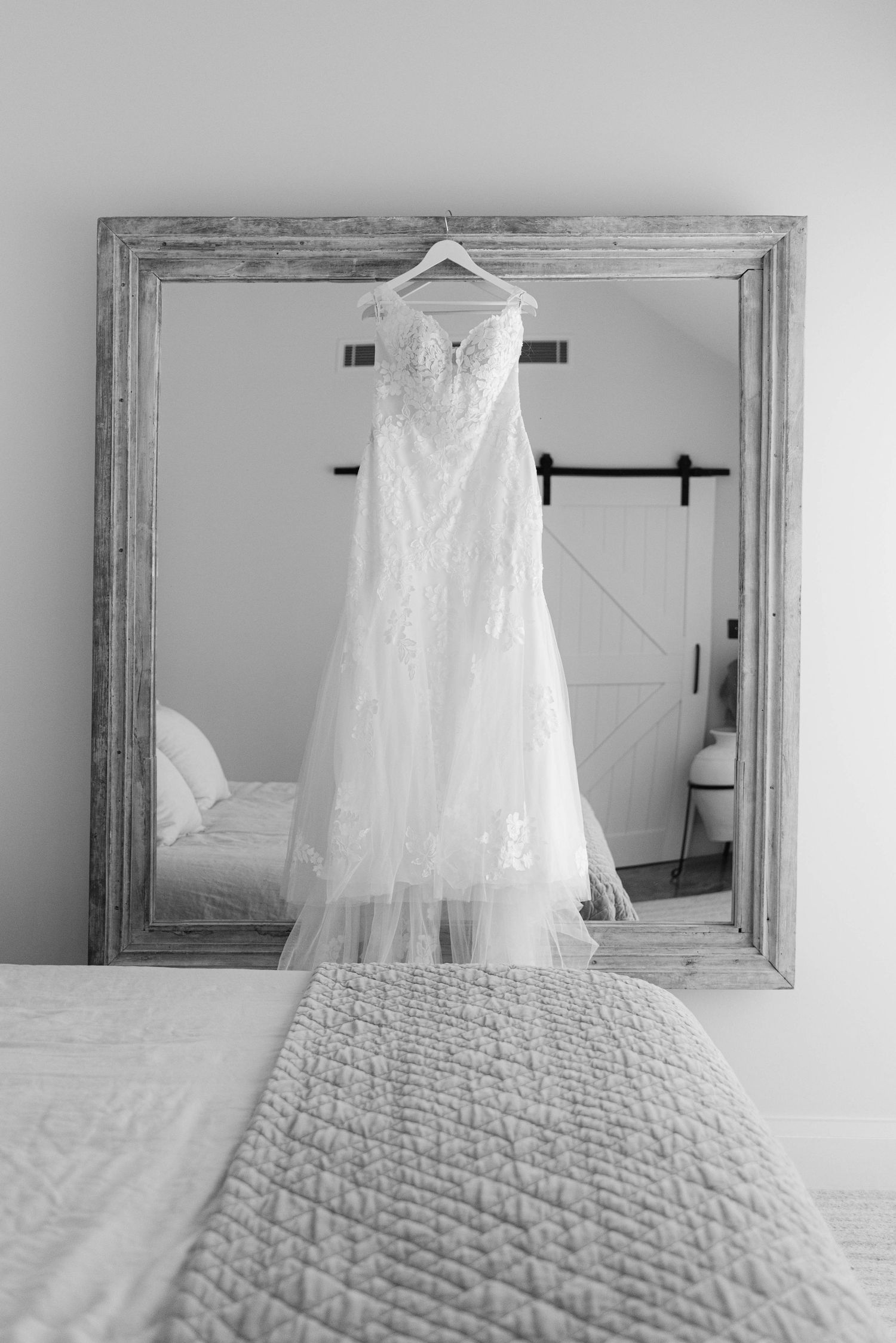 A white lace wedding dress hangs elegantly against a rustic mirror and textured bedding.