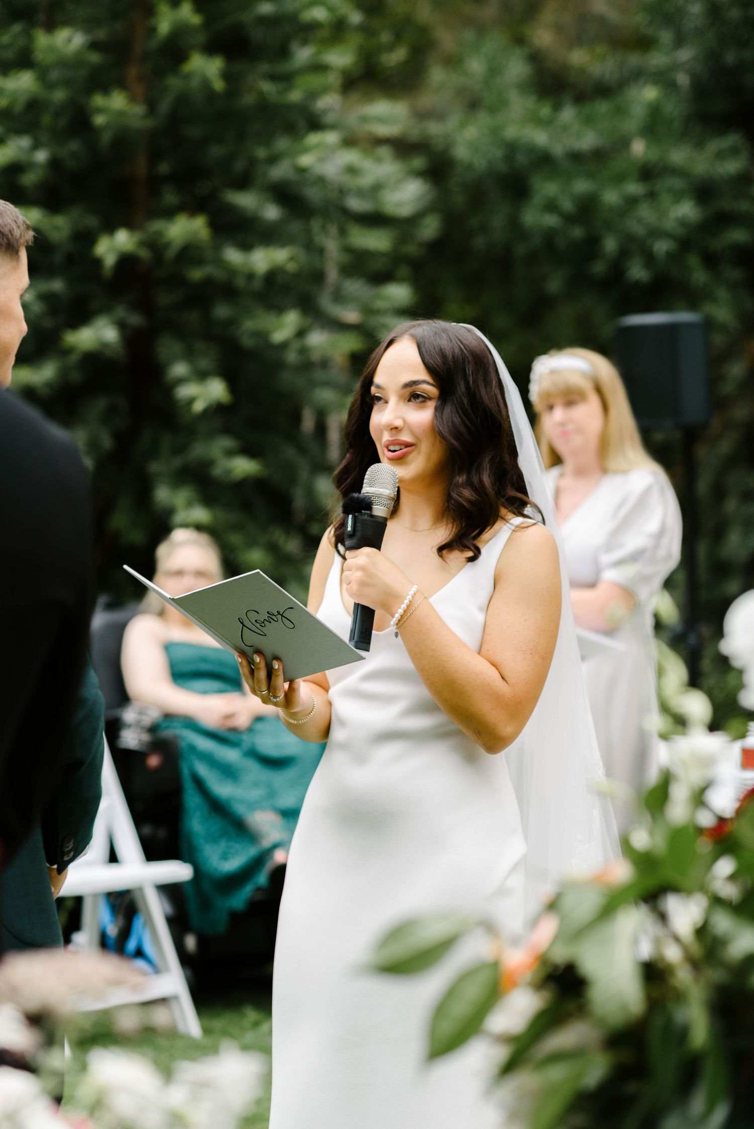 A bridesmaid in a white dress gives an emotional speech at an outdoor wedding reception while holding a paper.