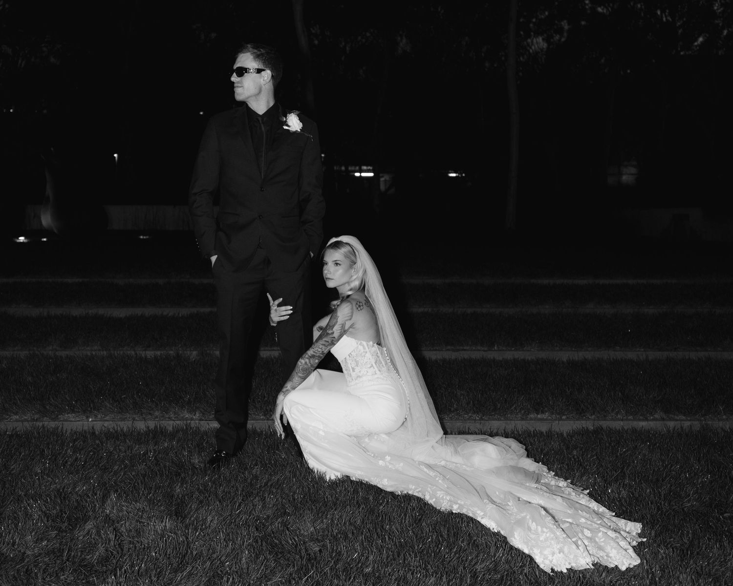 Black and white wedding photo of bride in gown seated on grass beside groom in suit and sunglasses at night.