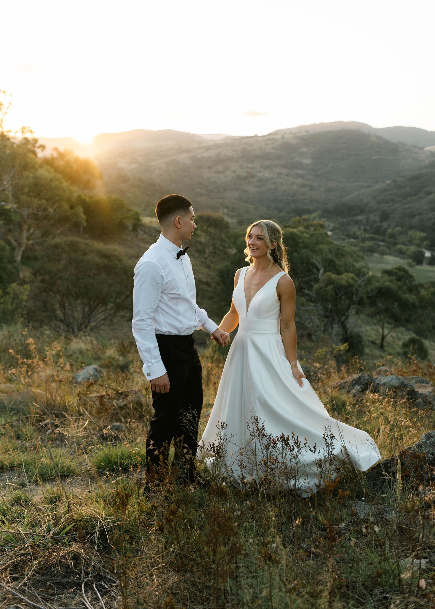A couple in wedding attire stands on a hillside during golden hour sunset with mountains in the background.