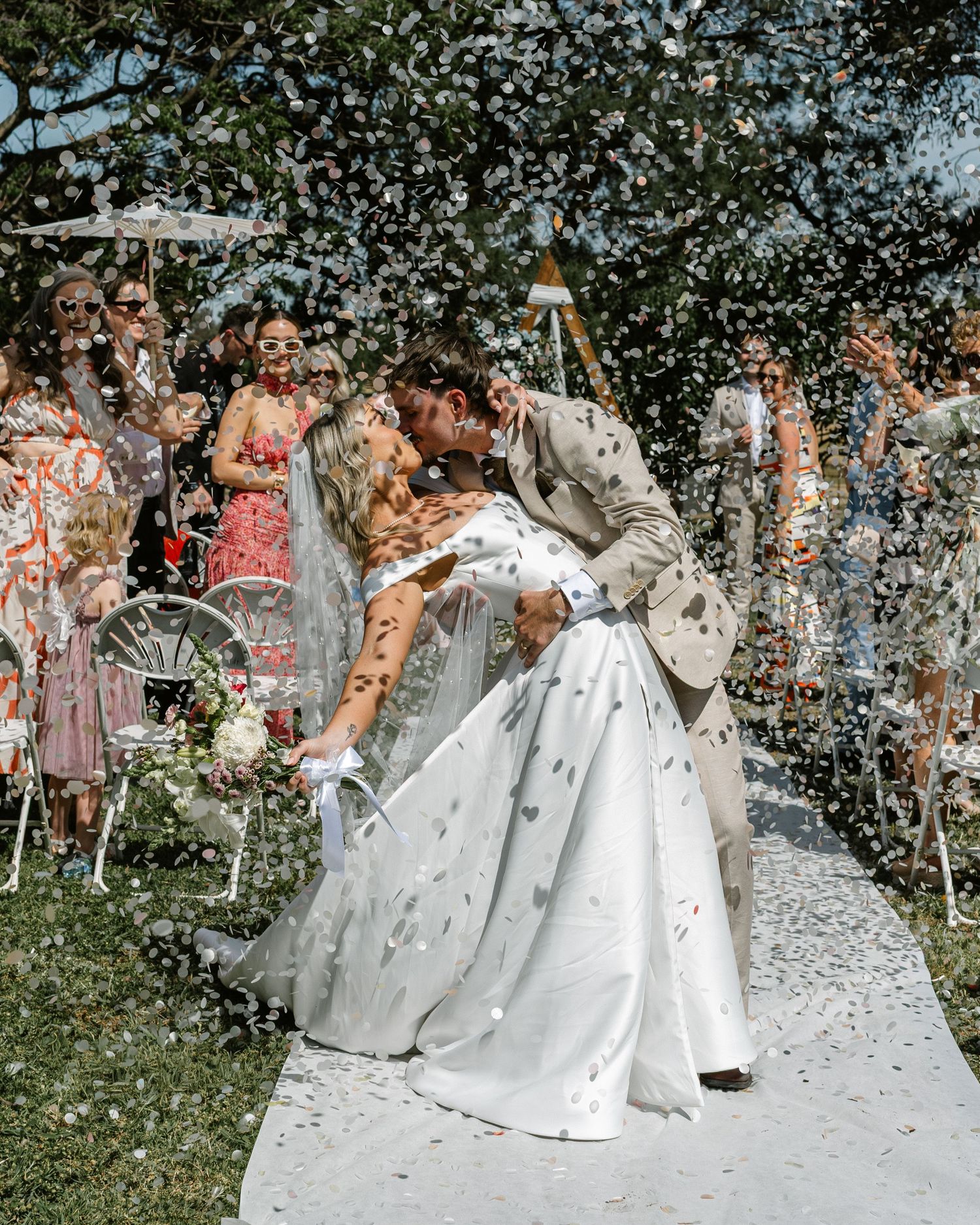 Bride and groom share a romantic dip kiss amid confetti shower, surrounded by cheering guests at outdoor wedding ceremony.