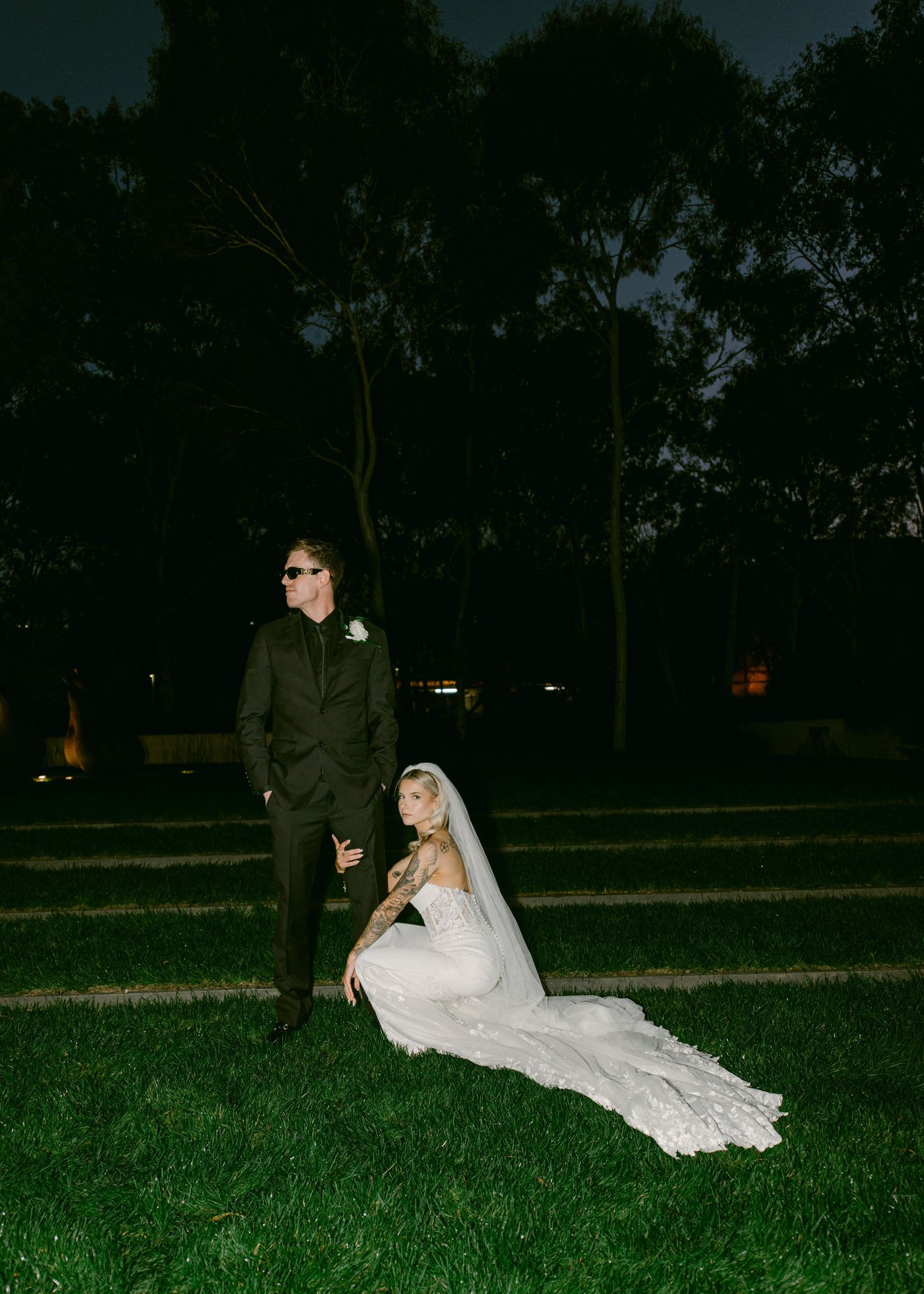 Bride in white lace gown and veil sits on grass while groom in dark suit stands beside her at night.