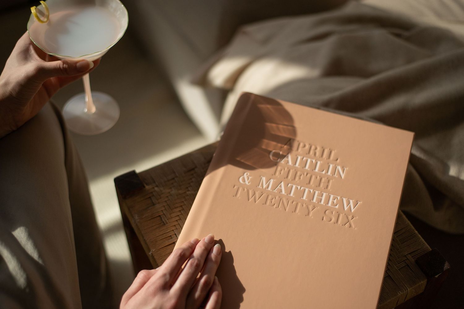 Elegant personalized wedding book with gold embossed names held by hands, with a cocktail glass in soft warm light.