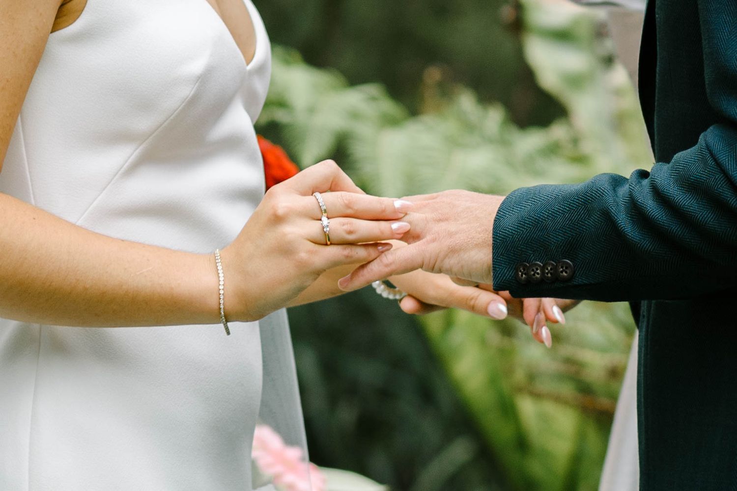 Close-up view of hands exchanging wedding rings during an outdoor ceremony with green foliage in background.