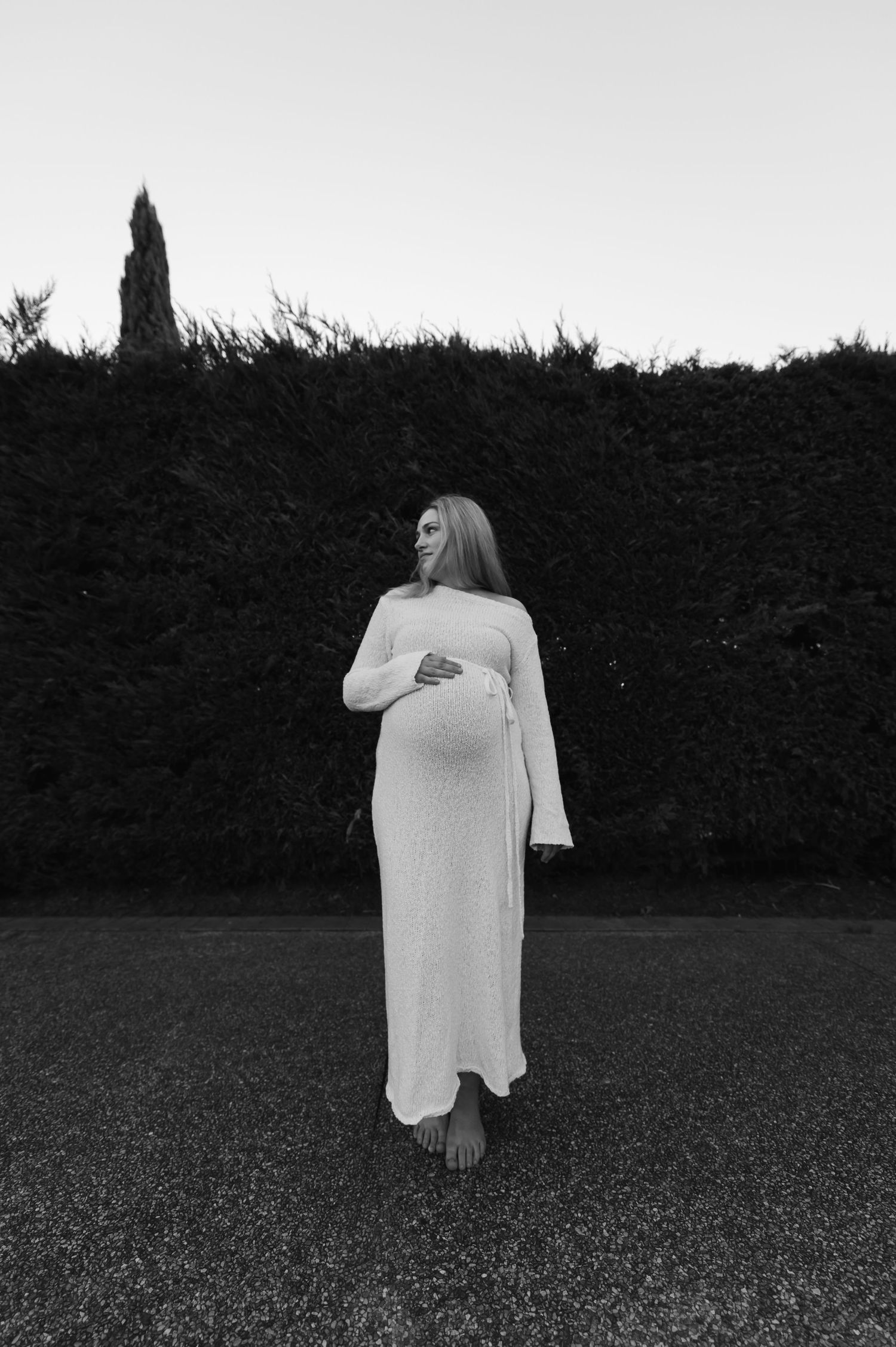 Black and white maternity photo of pregnant woman in white dress holding baby bump outdoors near hedge.