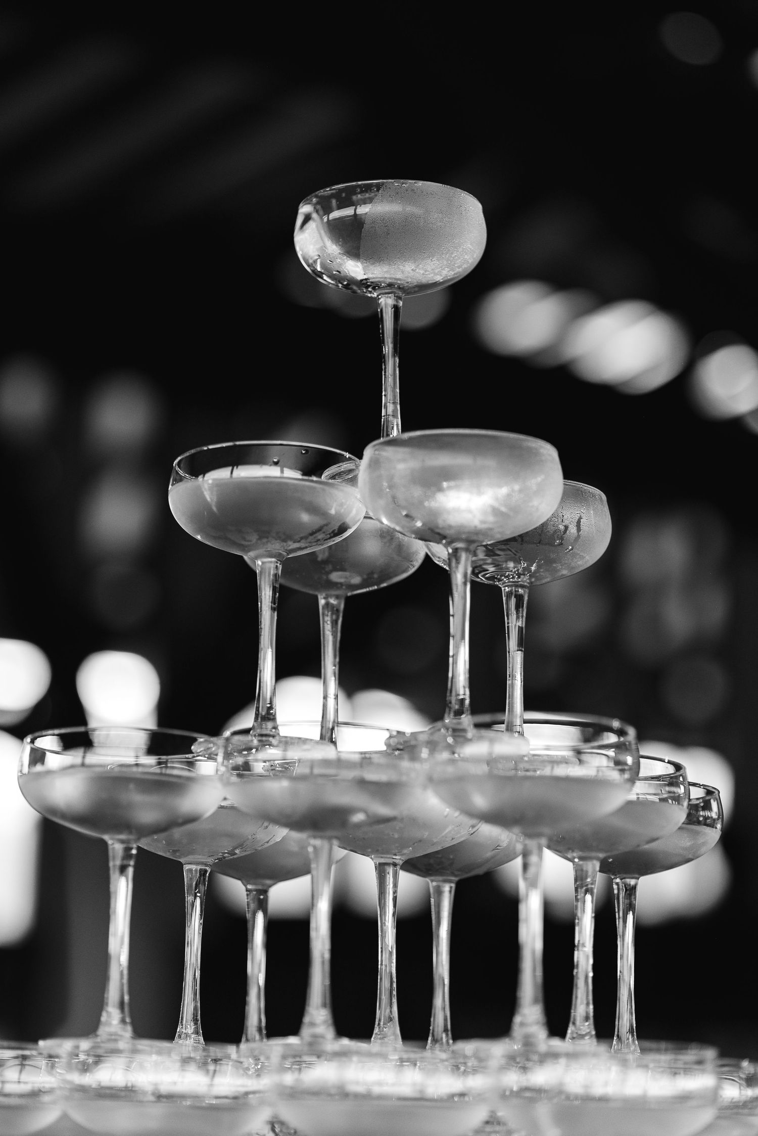 A black and white photograph of champagne glasses stacked in a pyramid formation with soft bokeh lighting effects.