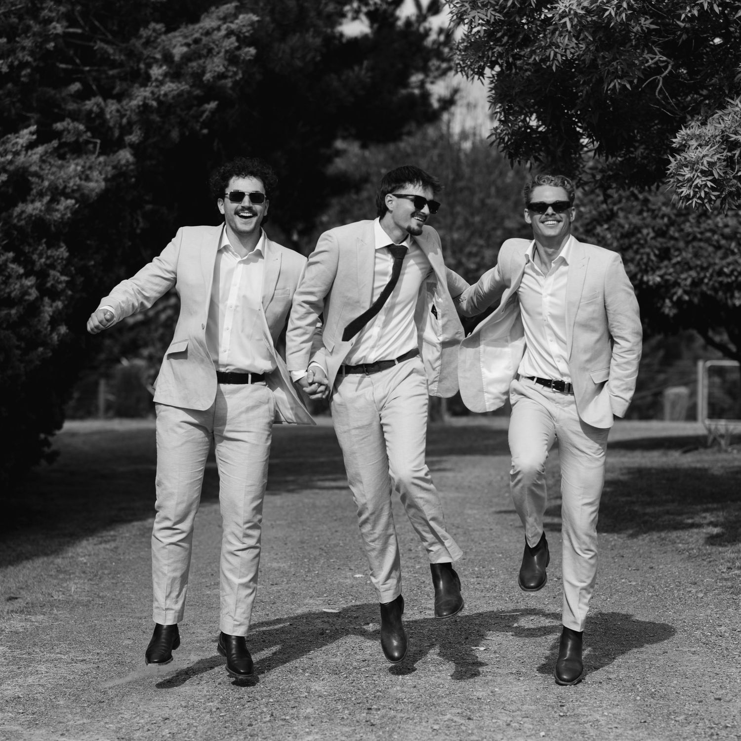 Groomsmen in light colored suits running and jumping playfully on a grassy path.