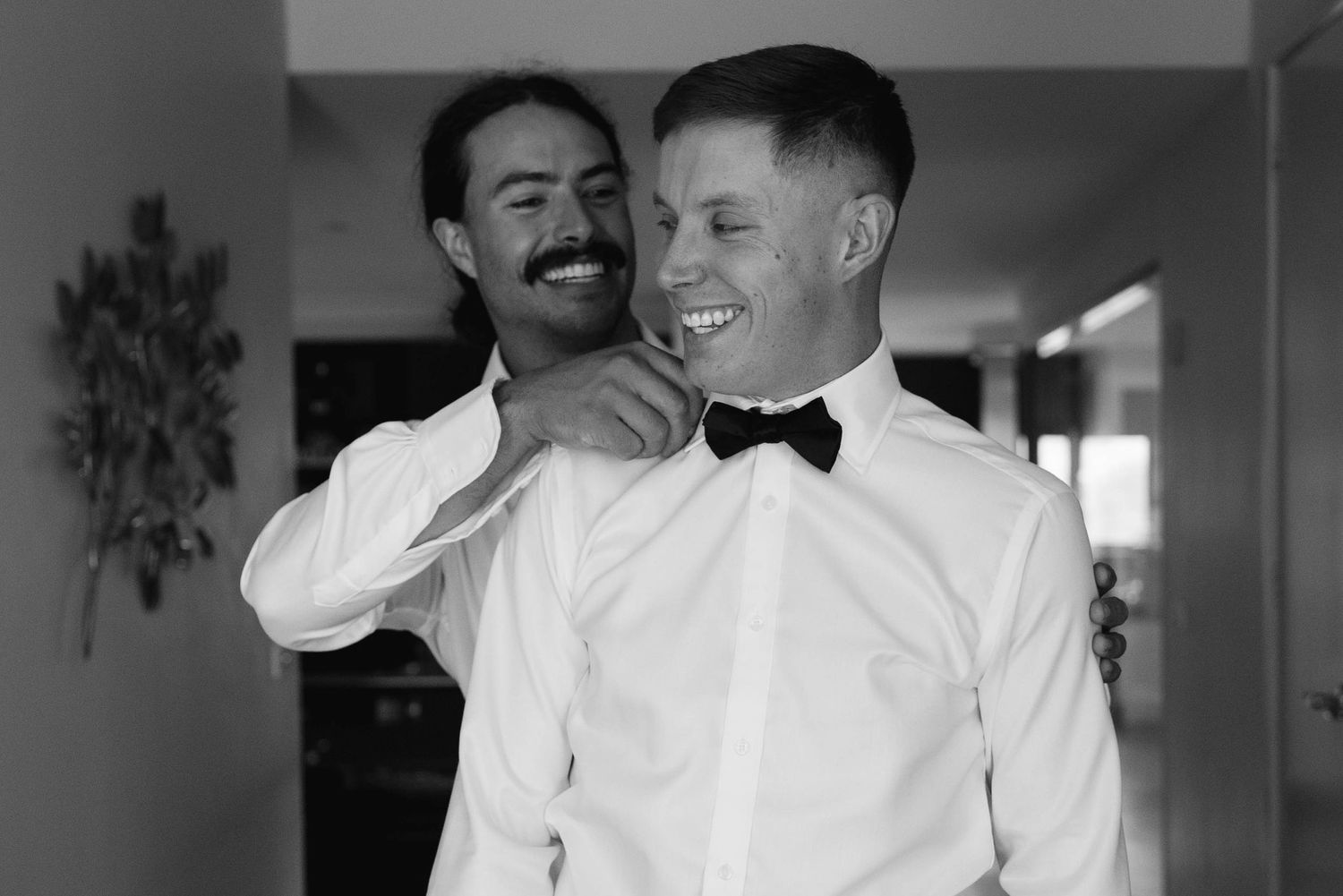 A group of groomsmen in white dress shirts and black bowties laugh together during a wedding celebration.