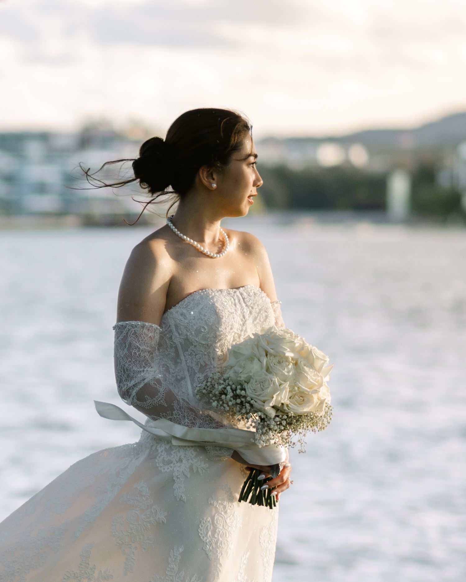 Bride in strapless lace gown holding white bouquet stands by waterfront, pearl necklace, hair up, golden hour light.
