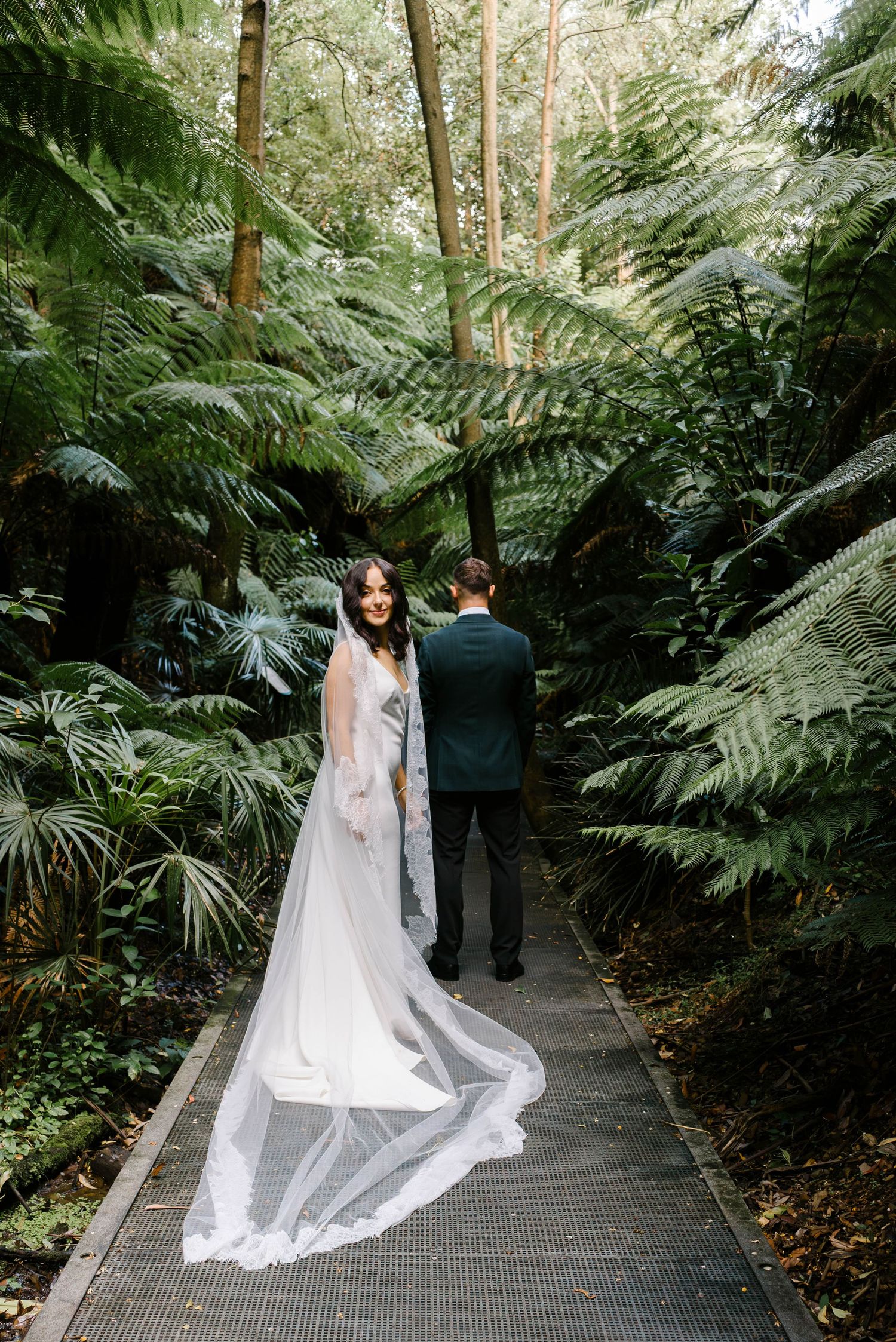A couple in wedding attire stands on a wooden boardwalk surrounded by lush green ferns and tropical foliage in a forest setting.