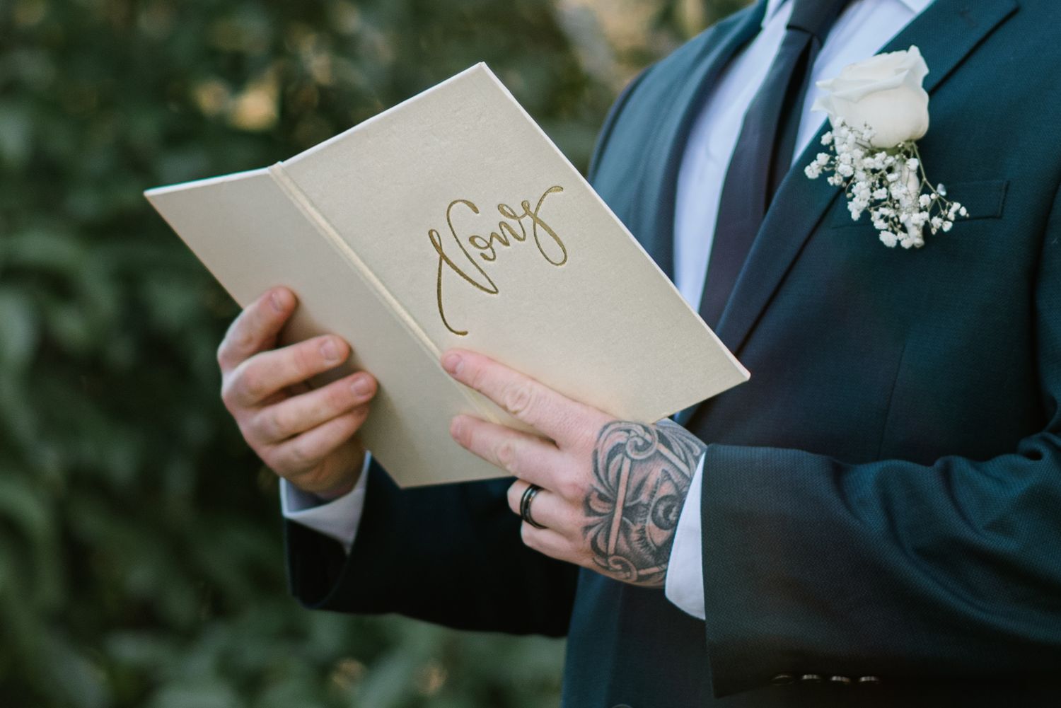 Groom in navy suit reads vows from elegant cream booklet with cursive script, adorned with white boutonniere.