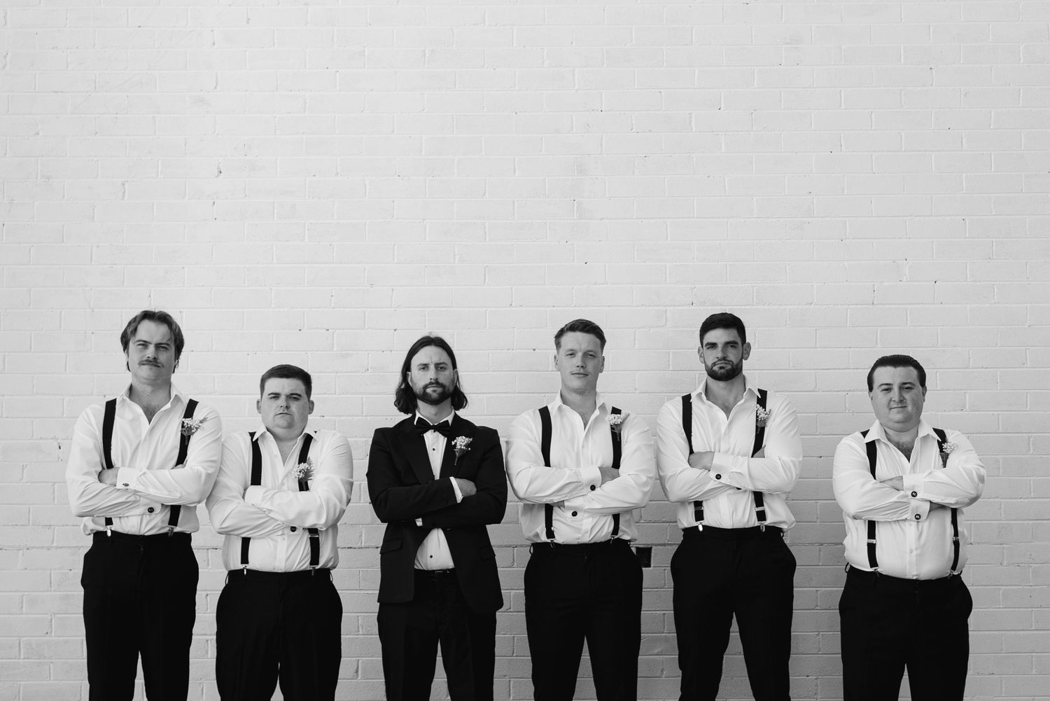 A lineup of groomsmen in matching white shirts and suspenders posing against a white wall.