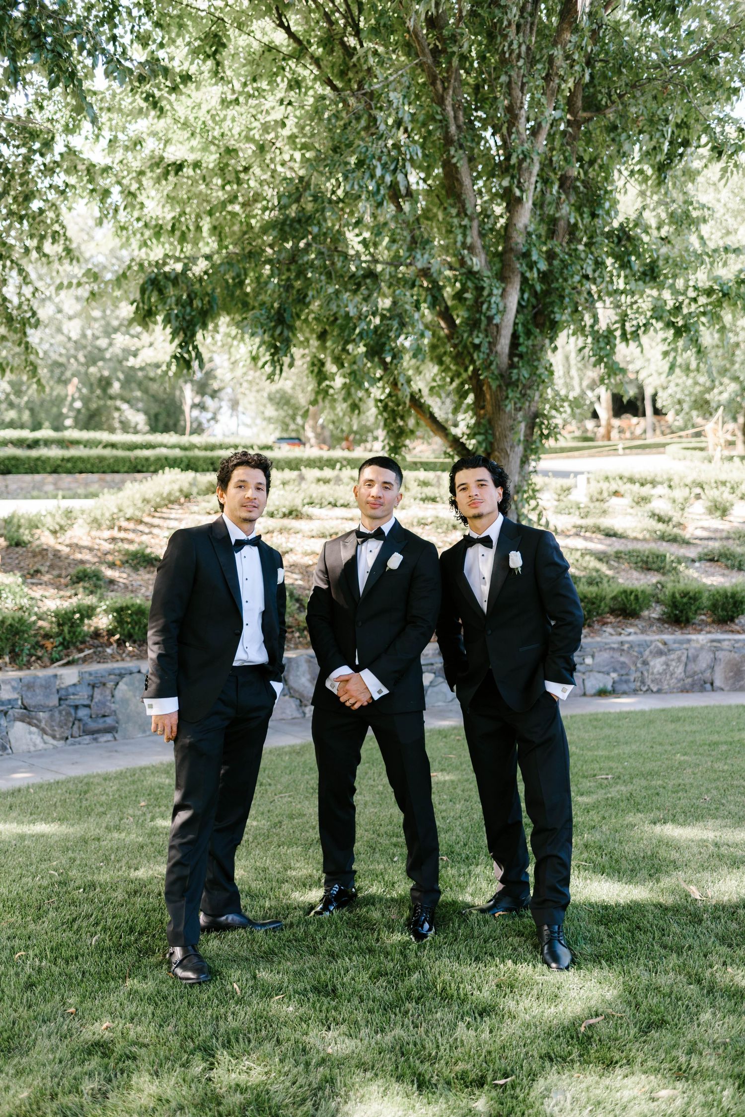 Three groomsmen in black tuxedos with bow ties pose outdoors on green lawn with trees and garden backdrop.
