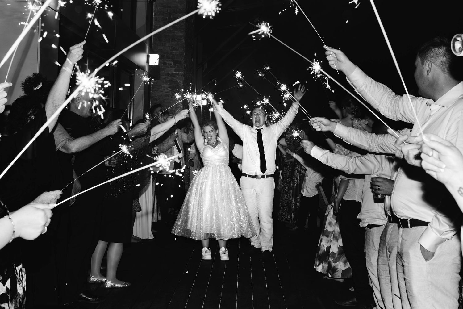 Wedding couple exits their reception under a tunnel of sparklers held by guests in black and white photograph.