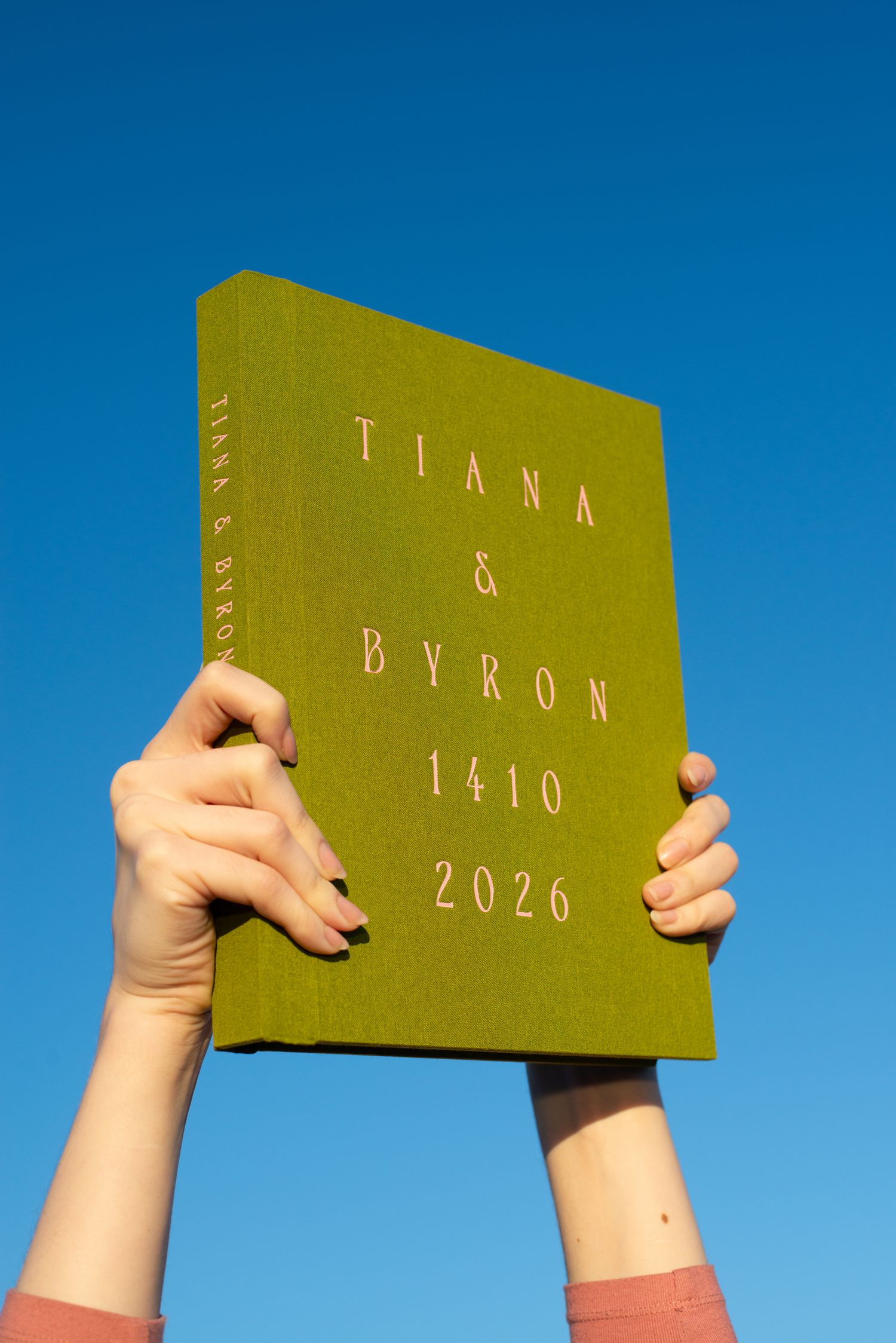 Olive green hardcover book titled Tiana & Byron 1410 2026 held up against a clear blue sky.